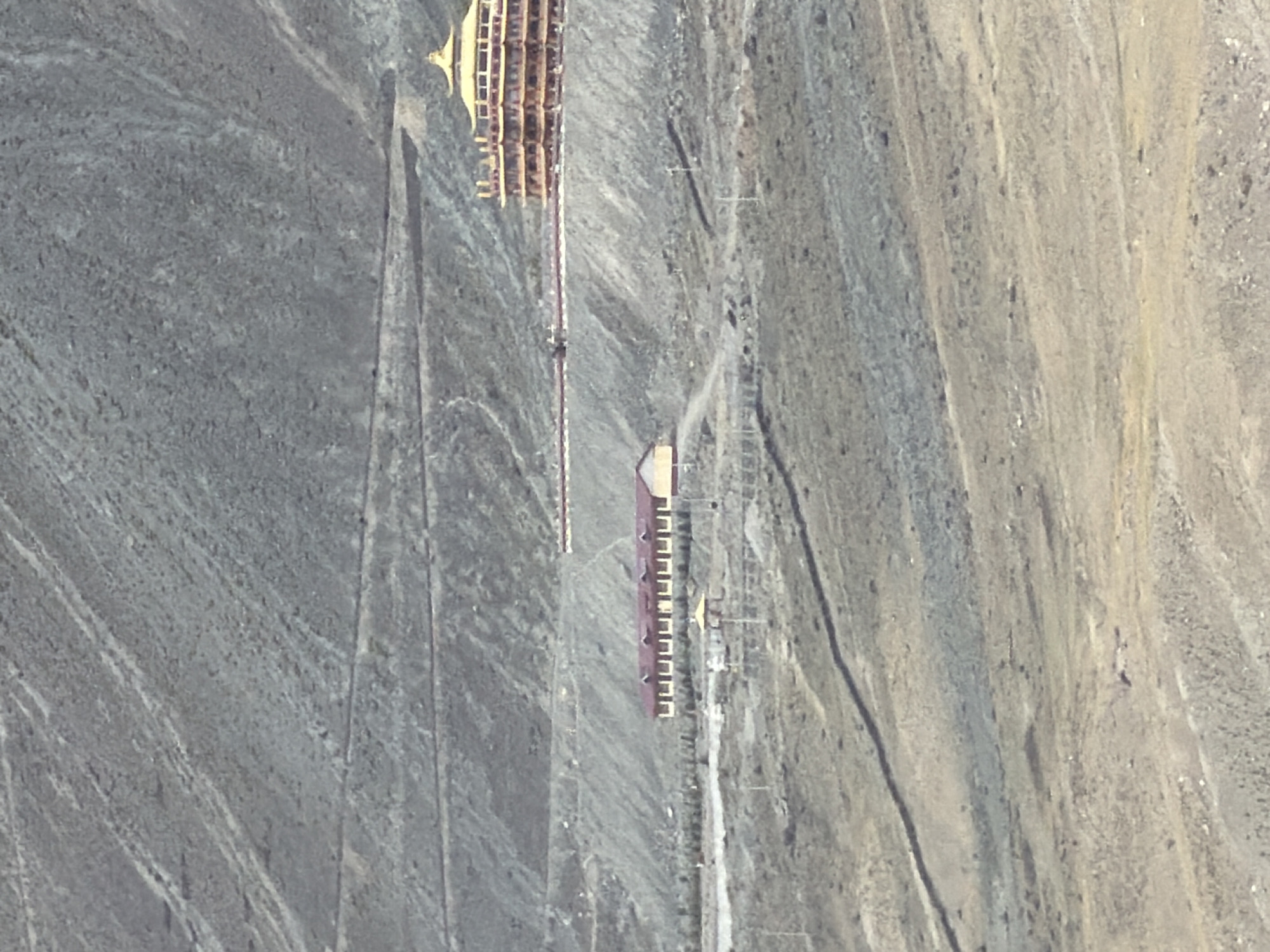 The image depicts a landscape with a winding road leading up a mountainside. A multi-story building with a golden roof, possibly a temple or monastery, is situated on the slope. Below this, another building with a red roof and yellow walls is visible. The environment appears to be arid and mountainous, with sparse vegetation. The overall scene suggests a remote, possibly high-altitude location.