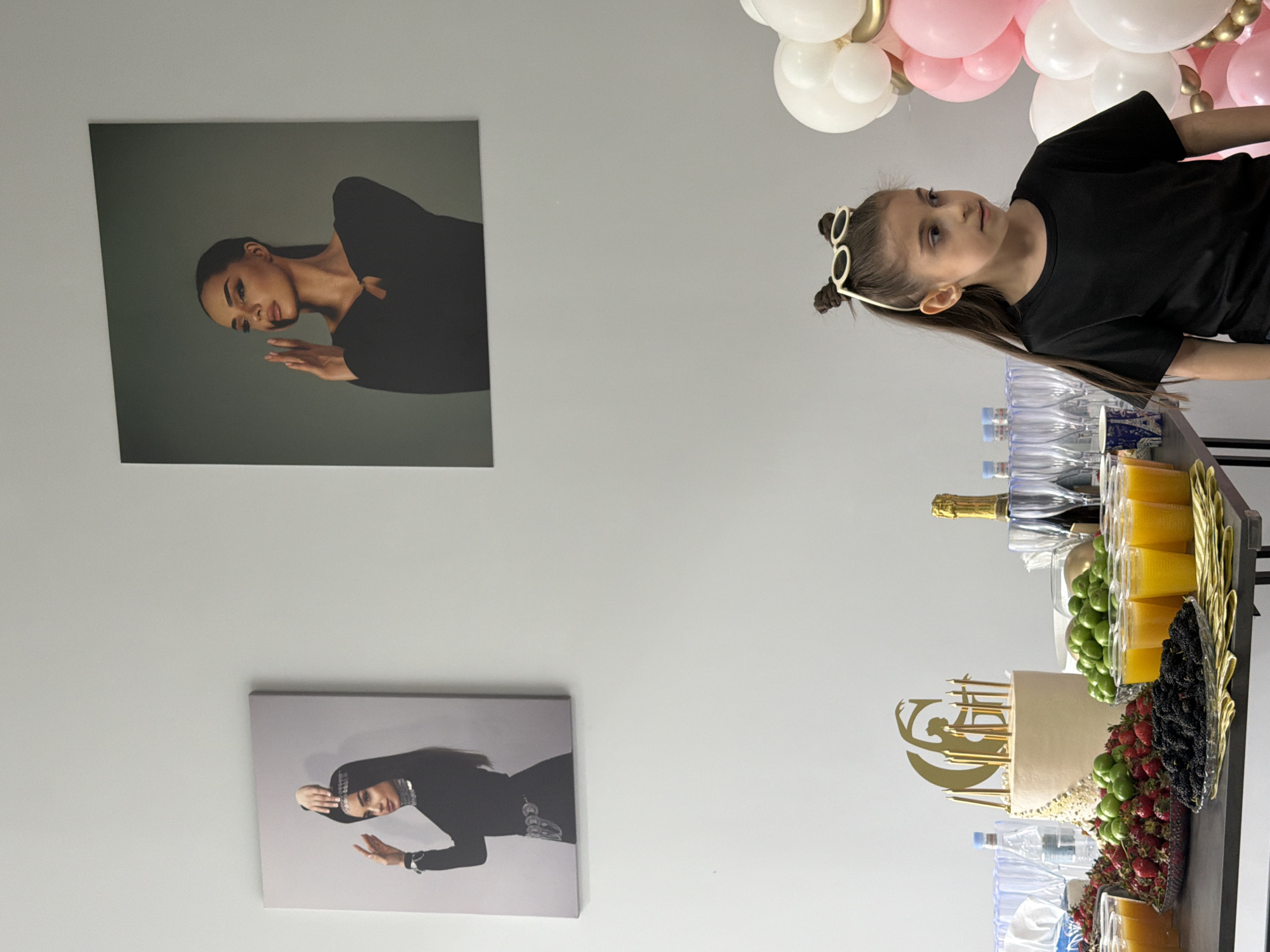A horizontal shot captures a child in a black shirt standing near a party table. The child, whose hair is styled with two buns adorned with sunglasses, is looking towards the left of the frame.
The party table is stocked with drinks, including bottled water, champagne, and juice. In addition, there is a cake with golden decorations and fresh fruits. Balloons in pink and white hues are positioned at the top right corner of the image, suggesting a celebratory atmosphere.
On the wall to the left of the child, two framed posters featuring women in black attire are visible. The location is a room with white walls. The city of Yerevan, Armenia is not visibly identifiable in the image.