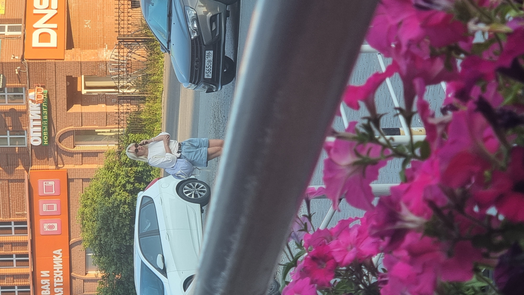 The image depicts a street scene in front of commercial buildings in Megion, Russia. A woman with blonde hair is standing on the sidewalk, talking on a mobile phone and carrying a light blue backpack. She is wearing a white t-shirt and denim shorts. Behind her, a white hatchback car is parked. To the right, a dark-colored car is visible, with a license plate reading "P565OB 186". The buildings in the background feature signage for "ОПТИКА" (Optics) and "DNS", indicating retail establishments. Pink petunias are in the foreground, partially obscuring the view. The lighting suggests it is daytime.