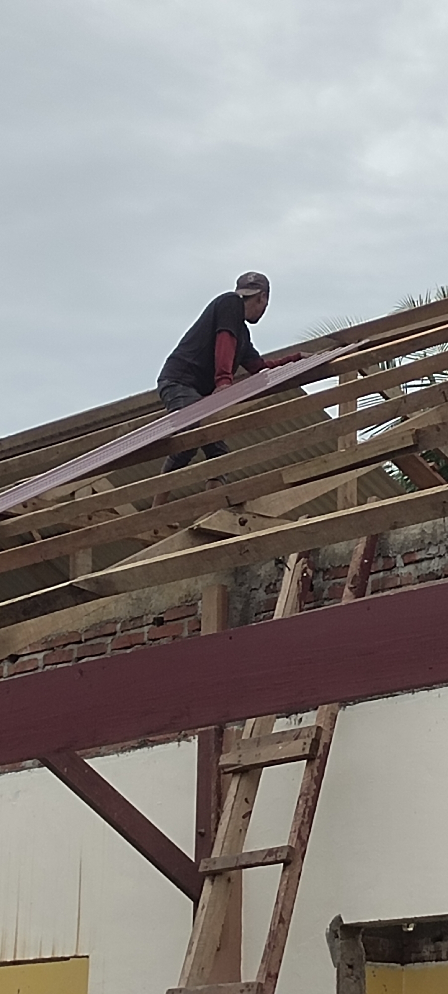 A man in a dark shirt and red gloves is working on the wooden frame of a roof in Langnibong, Indonesia. He is on the roof, placing a long, pink panel. The roof framing is incomplete, exposing brickwork. A wooden ladder leans against the structure. The sky is overcast.