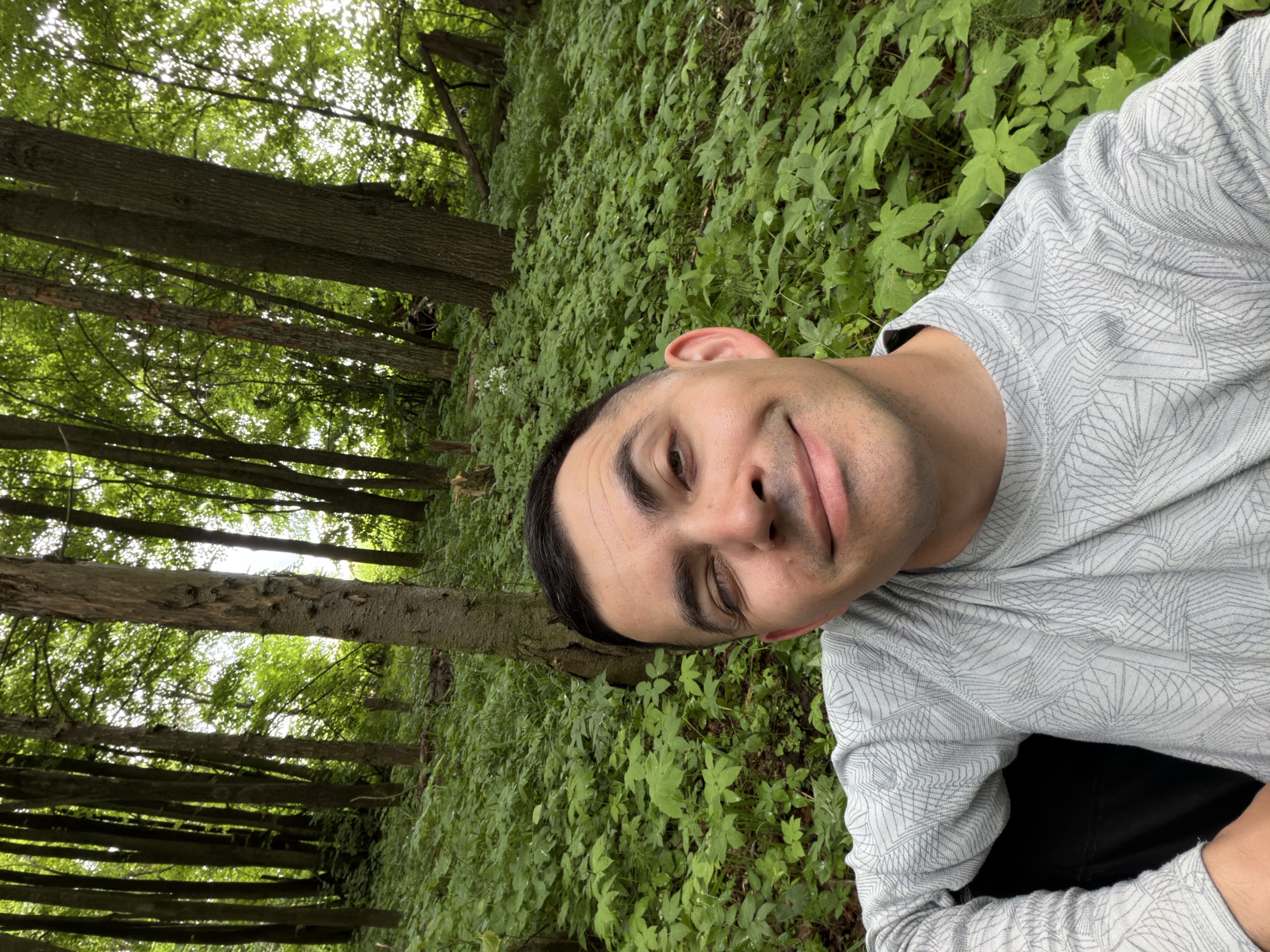 This image depicts an adult male in a close-up, selfie-style photograph taken in a wooded area in Kechur, Russia. The man has short, dark hair and dark eyebrows, with a light-to-medium skin tone, and is captured with a slight smile, looking directly into the camera. He is wearing a light grey long-sleeved shirt featuring a subtle, repeating geometric pattern of interconnected lines. A dark, indistinct object, possibly a bag or clothing, is partially visible behind his left arm and shoulder. The background consists of a dense forest environment with numerous tall trees, exhibiting various bark textures and hues. The ground is thickly covered with a uniform layer of bright green undergrowth, some leaves appearing to have water droplets. The lighting is diffused, suggesting an overcast sky or light filtering through a dense tree canopy. No other individuals or man-made structures are visible.