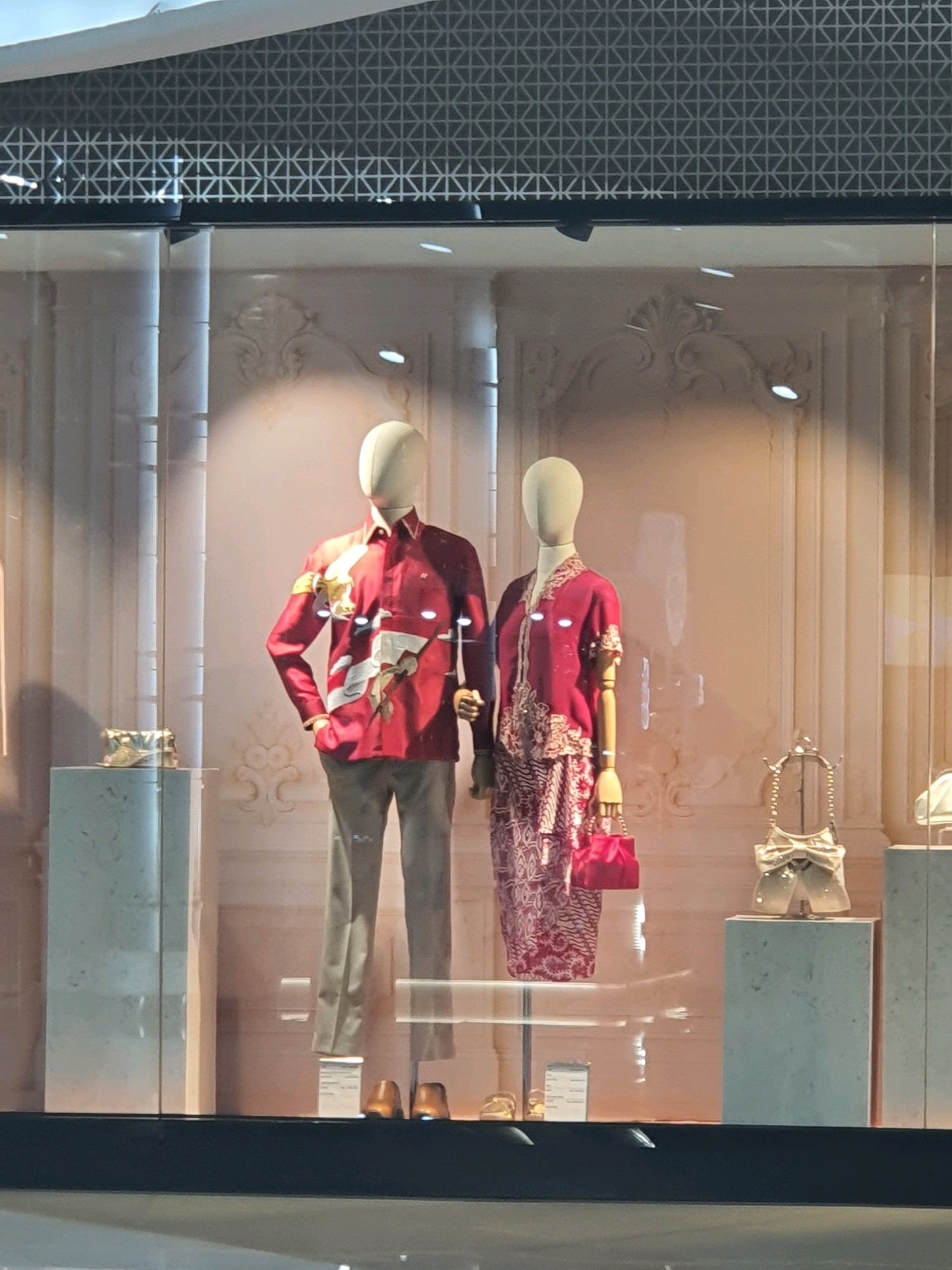The image captures a retail display window, likely in a shopping mall, featuring two mannequins. The mannequin on the left is male, dressed in a vibrant red long-sleeved shirt with a white and gold graphic print, paired with light gray trousers and brown shoes. The mannequin on the right is female, wearing a matching red ensemble with intricate white detailing, and holding a bright pink handbag. Both mannequins have featureless, light-colored heads.
The display is set against a backdrop of pale pink walls with ornate decorative moldings. To the left of the mannequins, a cream-colored rectangular pedestal displays a small metallic handbag. To the right, another similar pedestal showcases a decorative object resembling a bow with a chain. In the background, vertical fluted columns and horizontal decorative elements contribute to a sophisticated aesthetic. The lighting appears to be artificial, with several spotlights illuminating the display.
The scene is viewed from outside the display window, with reflections of the external environment visible on the glass. The top of the image shows a textured, geometric pattern on the ceiling or upper structure of the building. The foreground is a dark surface, possibly the floor outside the display or a ledge.
Based on the context provided, this display is located in North Jakarta, Indonesia.