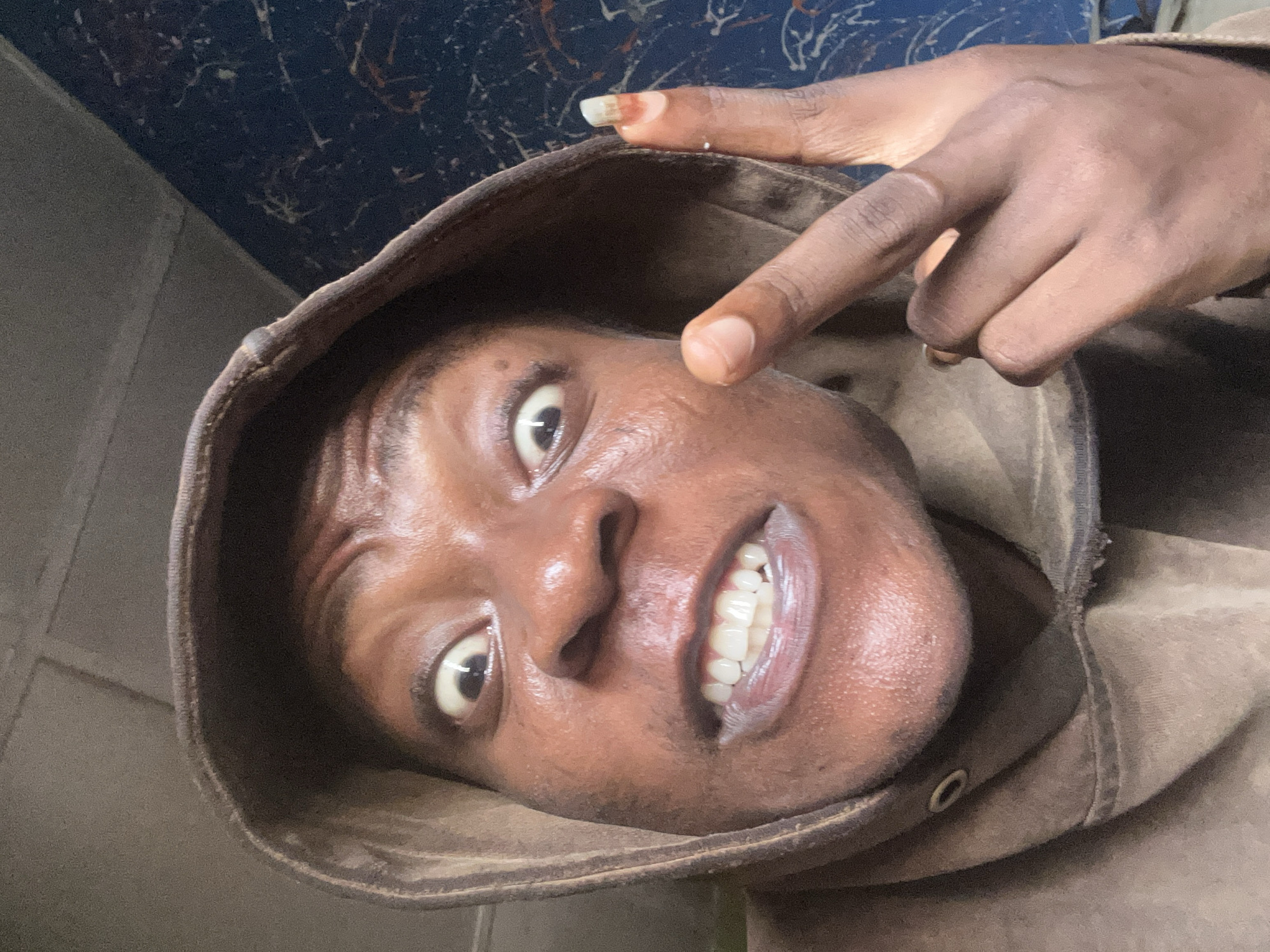 A close-up, tilted selfie of a person with dark skin, wearing a brown hooded sweatshirt. The person is pointing a finger towards their face, displaying white teeth in a wide smile. The background is obscured but appears to be a wall of dark blue with speckled white markings and a grey wall. The location is unknown.