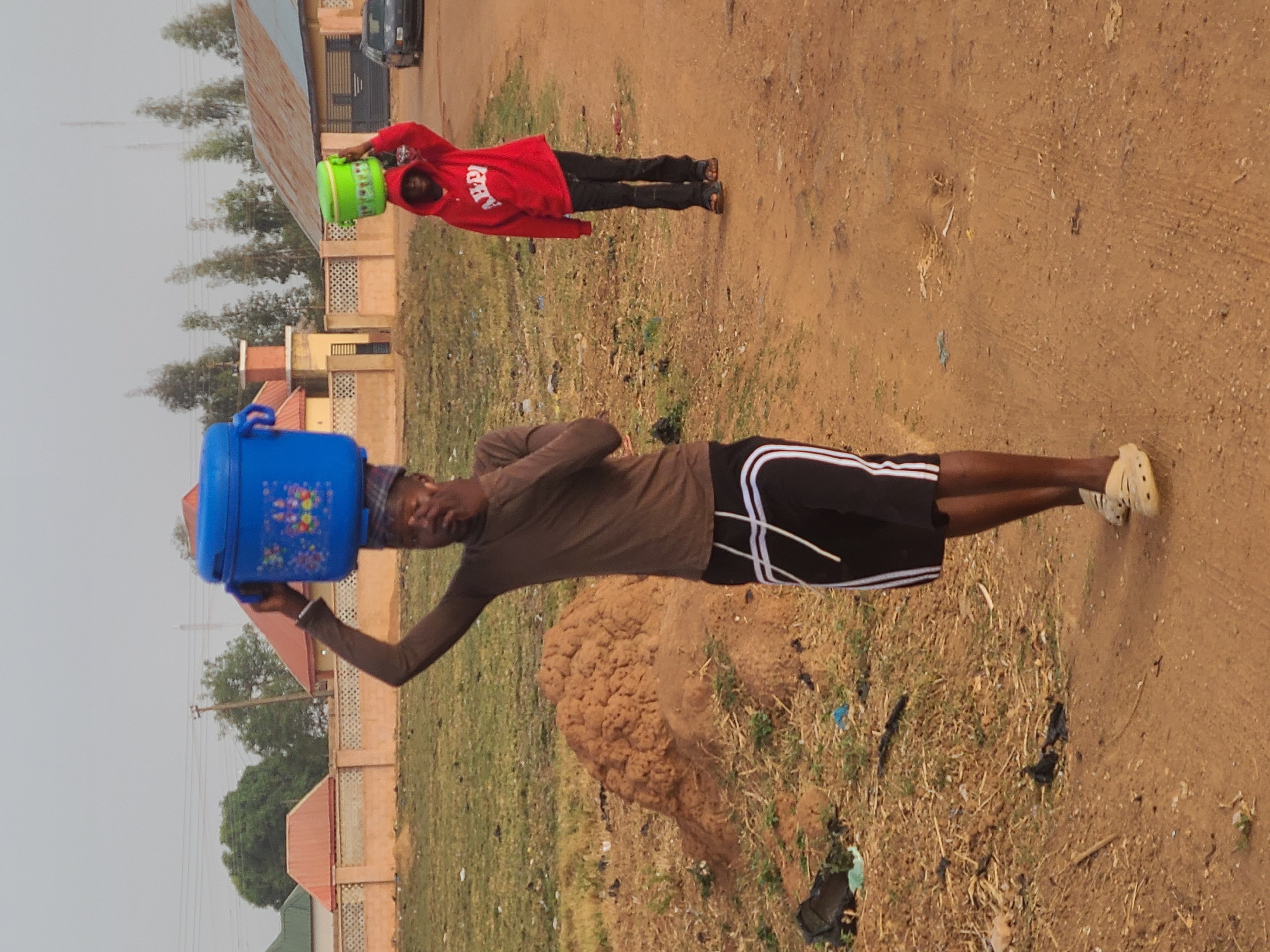 The image depicts two individuals in a rural area of Barakin Lerawi, Nigeria. One person, wearing a brown shirt and black shorts with white stripes, is carrying a blue plastic container on their head. The container is decorated with floral patterns. The person is holding the container with their right hand. The second person, further away, is wearing a red hooded jacket and black pants, also carrying a green container on their head. Both individuals appear to be walking on a dirt road. In the background are buildings and sparse vegetation. The ground is mostly dirt with patches of grass and debris.