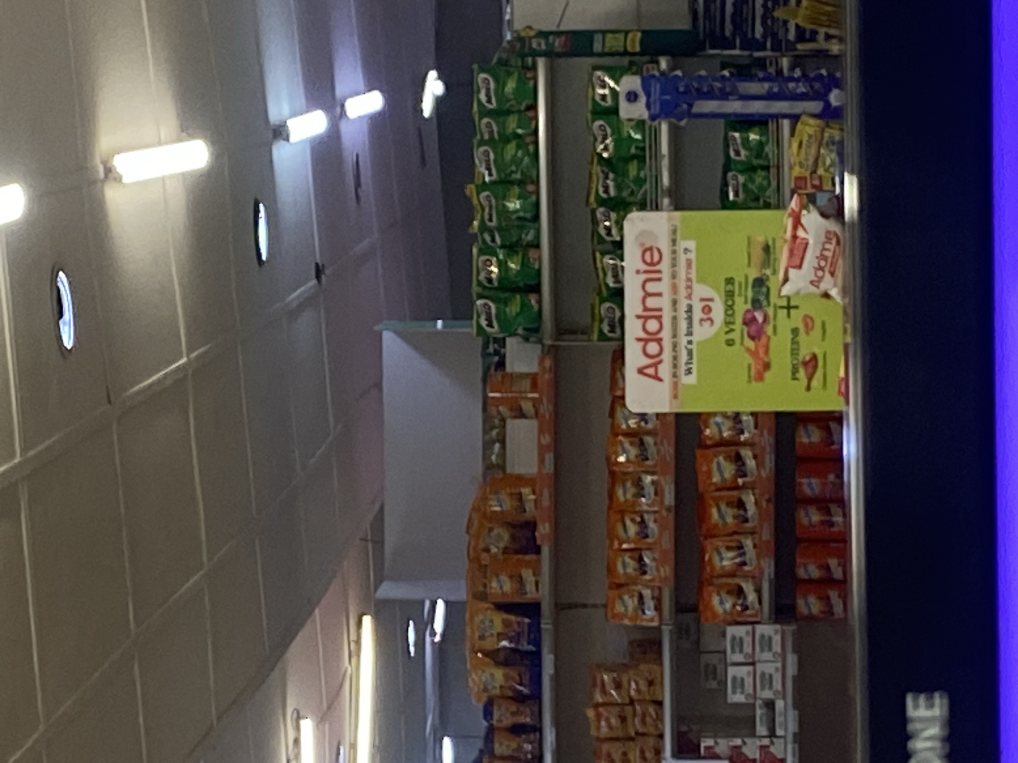 This media file captures an indoor view of a retail setting, likely a supermarket or convenience store, in Ikorodu, Nigeria. The scene is dominated by shelves stocked with various food items and other products. In the foreground, a prominent display features a green and yellow sign for "Addmie," a product that seems to contain "6 Veggies" and "Protein." The sign also asks, "What's inside Addmie?".
The shelves are filled with packaged goods, including what appear to be snack bags with green branding, possibly Milo, arranged in vertical rows. Further down, orange-packaged items are stacked, and smaller white boxes are visible near the bottom. The lighting suggests it is daytime, with bright fluorescent lights illuminating the aisles. The overall impression is one of a well-stocked retail environment, with products neatly displayed for shoppers. There are no people visible in the frame. The blue edge on the right side of the image might indicate the presence of a display screen or a structural element of the store. The visible text "ONE" on the far right edge of the image could be part of a store name or branding.