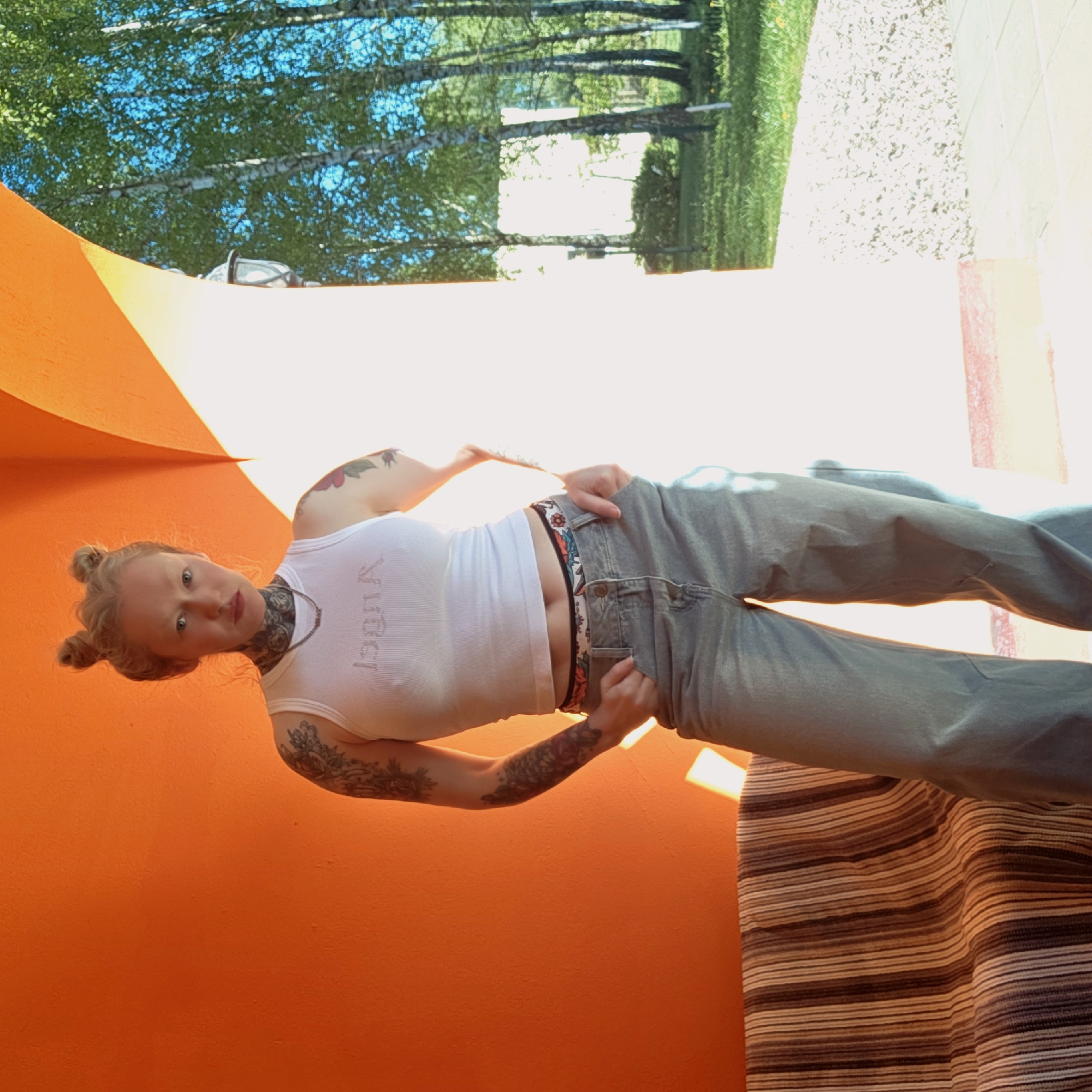 A woman with blonde hair styled in two buns is standing indoors in front of a bright orange wall. She is wearing a white tank top with text, light wash jeans, and a patterned belt. Her left arm and chest have tattoos. The location appears to be in Тэльмінскі сельскі Савет, Belarus, as evidenced by environmental features visible through a partially open doorway or window. These include trees, green vegetation, and a light-colored path or driveway. A striped rug or blanket is partially visible at the bottom right of the image.
