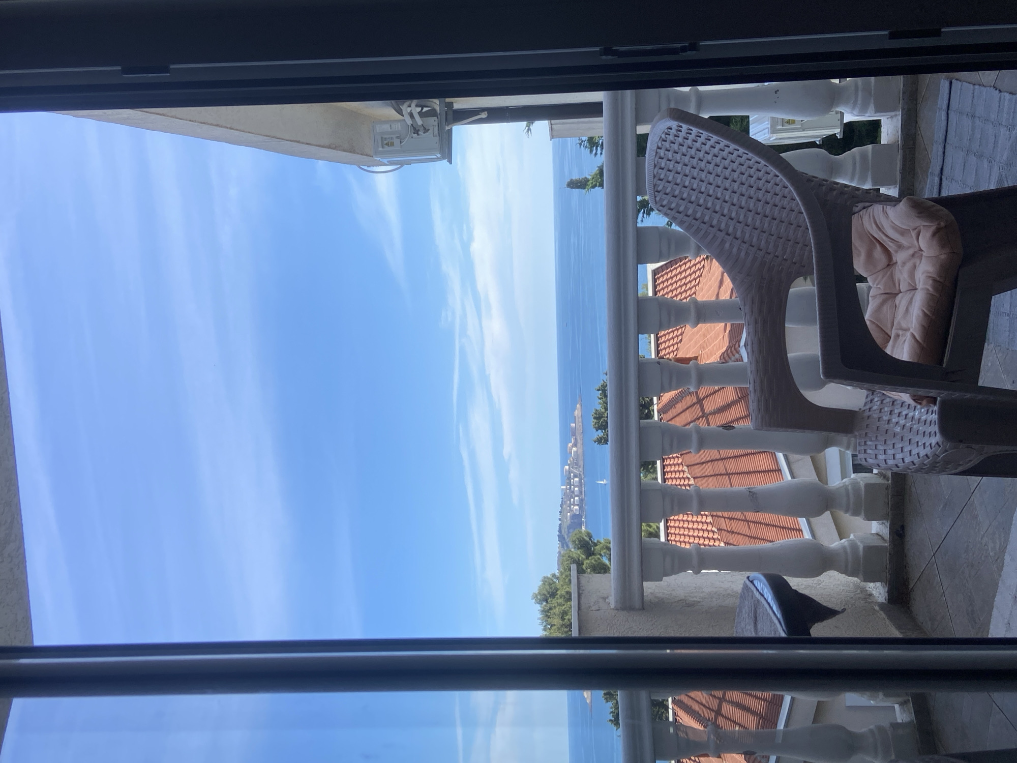 View from a balcony overlooking the Adriatic Sea, likely in Šušanj, Montenegro. A brown plastic chair with a beige cushion sits on the balcony. White balustrades line the balcony edge. Red tiled roofs of buildings are visible below. Across the sea, a coastal town is discernible. The sky is blue with light clouds. The image is framed by a window or doorway. An air conditioning unit is mounted to the exterior wall.