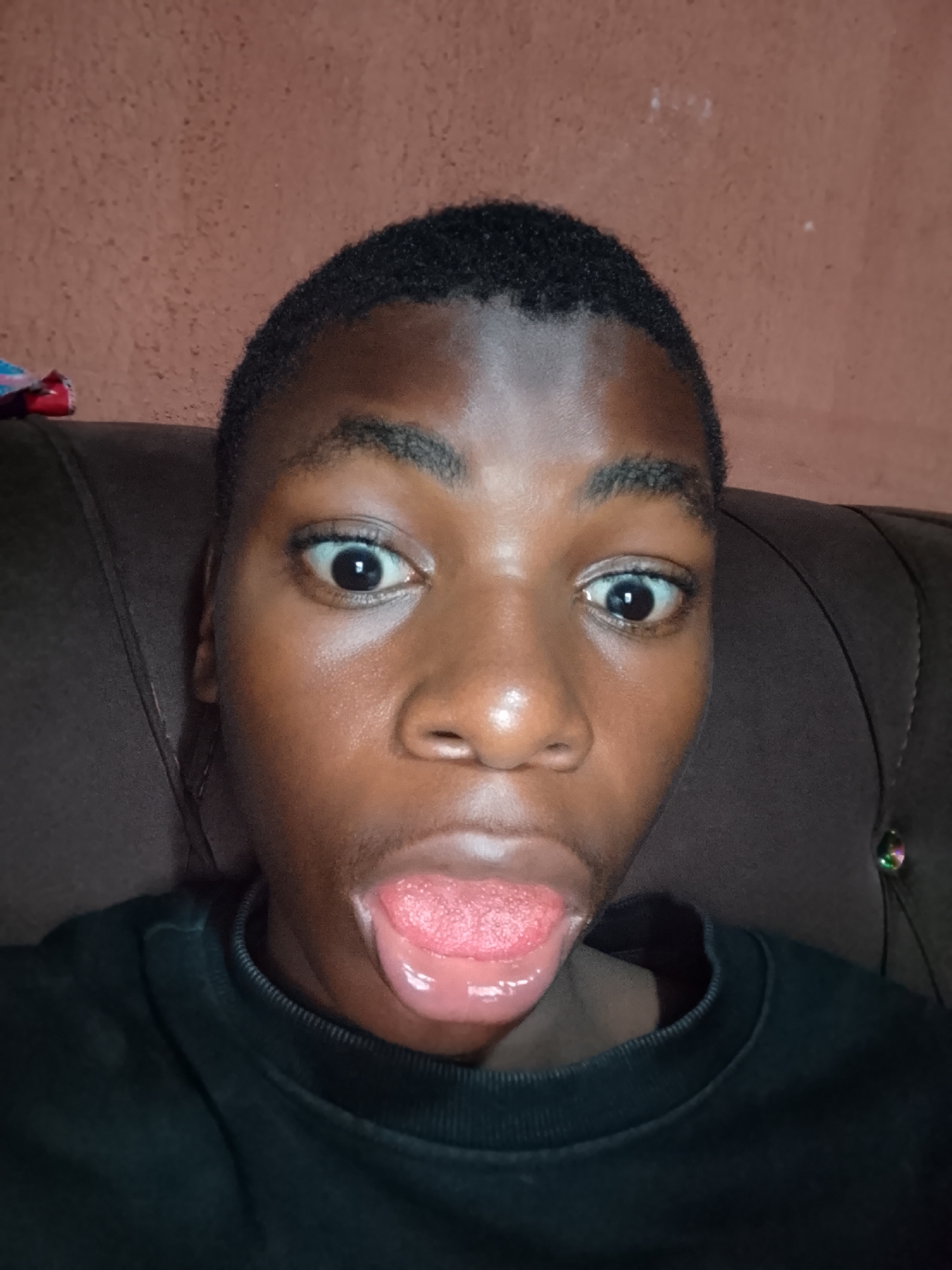 The image presents a close-up portrait of a young person with dark skin and short, dark hair, looking directly into the camera. Their eyes are wide open, and their tongue is extended in a playful or humorous expression. They are wearing a dark, possibly black or dark blue, crew-neck top.
The background shows an indoor setting. In the upper portion, a reddish-brown, textured wall is visible. Behind the person and extending to the sides is a dark brown, cushioned piece of furniture, likely a sofa or chair. A small, colorful object, possibly red and blue, can be seen on the far left against the wall. The lighting appears to be artificial, creating highlights on the person's forehead, nose, and the moist surface of their tongue. No text is visible in the image.