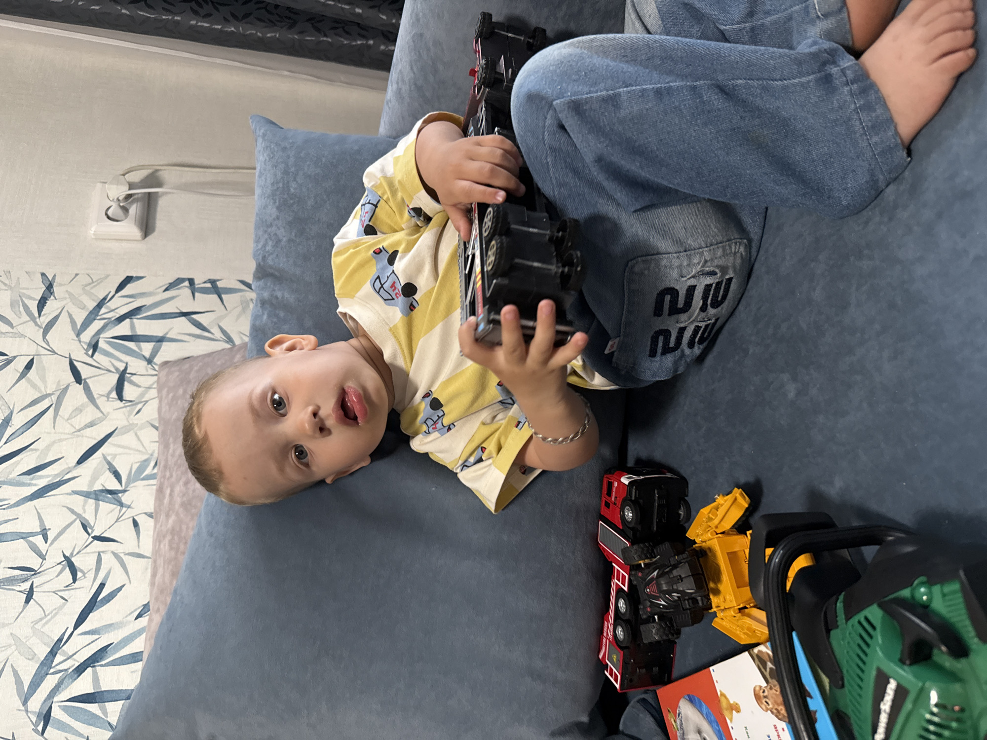 A young child, appearing to be a toddler, is lying on a blue upholstered surface, possibly a sofa or large cushion. The child is wearing a yellow and white striped t-shirt with car designs and blue jeans. The child is holding a black toy truck. Several other toys are scattered nearby, including a red fire truck, a yellow construction vehicle, and a green toy chainsaw. The background shows a wall with an electrical outlet and a decorative pattern of blue and white leaves. The location of Rusanovo, Russia is not discernible from the visual elements present in the image.