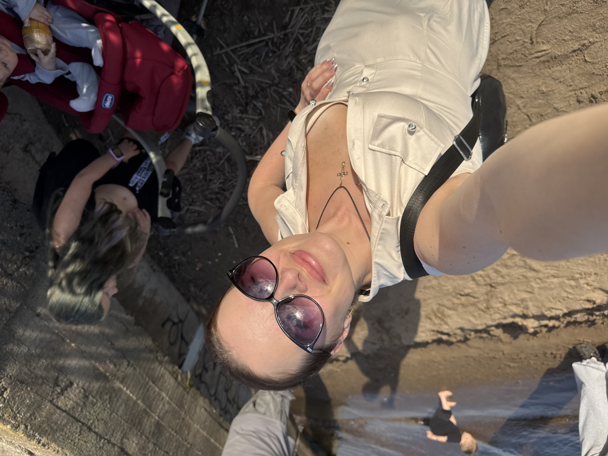 In a bright, outdoor scene on a sandy embankment, a woman in a light-colored dress and sunglasses takes a selfie. She wears a gold cross necklace. To her left, a black strap from a bag is visible. In the background, a child in a black shirt plays near the water's edge. To the right, a young girl with long, dark hair sits near a concrete structure, and a red stroller with a baby is visible. The environment suggests a park or a recreational area near a body of water. The setting appears to be the embankment of the Votkinsk Reservoir in Chaikovsky, Russia.