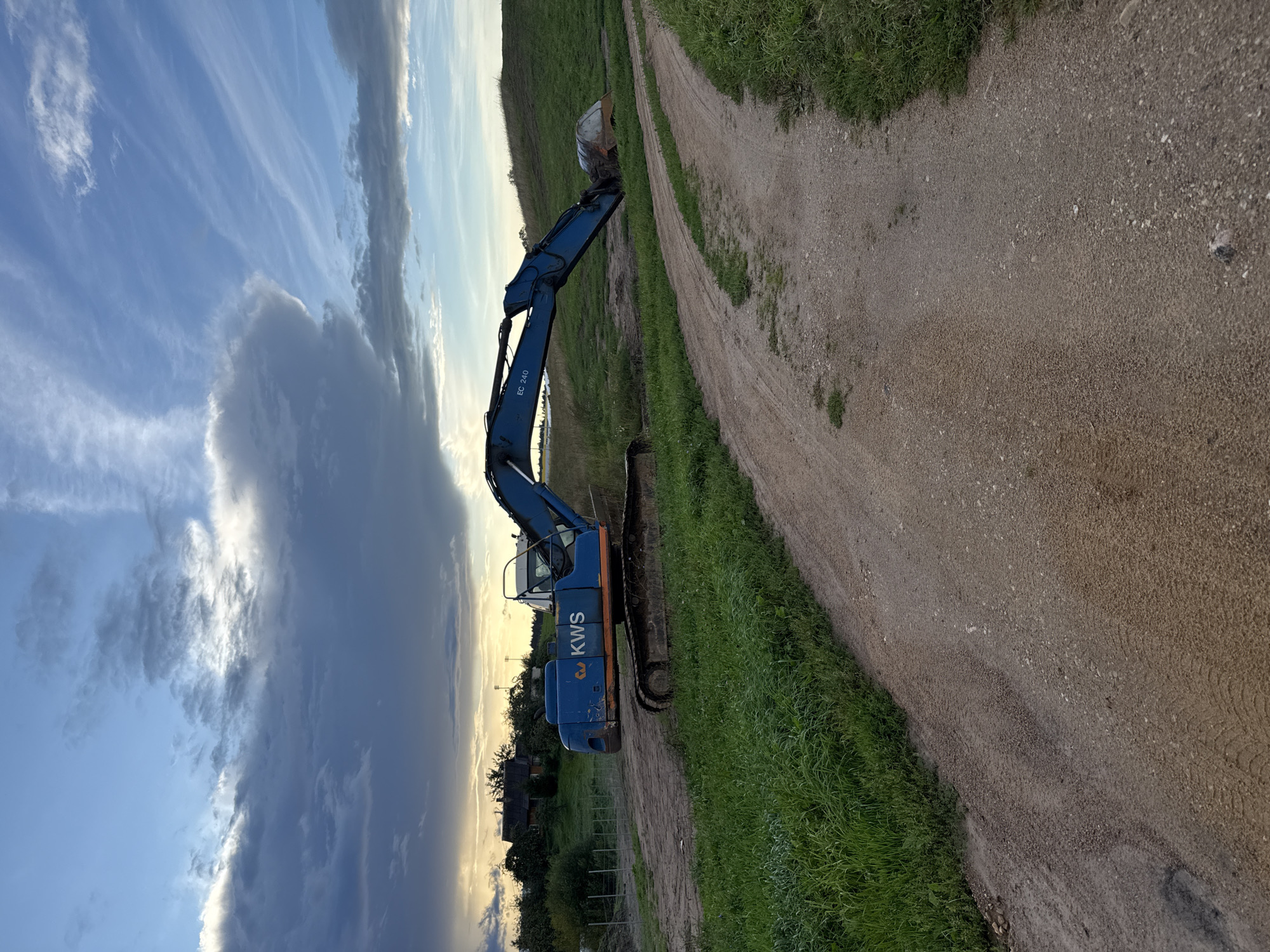 In the city of Galinė, Lithuania, a blue excavator with "KWS" branding sits idle on a dirt track next to a grassy field under a cloudy sky. The excavator's boom is extended, with its bucket partially submerged in a ditch. The scene appears to be an outdoor construction or excavation site, possibly in a rural or semi-rural area, with a few trees and a building visible in the background. The road in the foreground is unpaved and shows tire tracks, indicating recent vehicle activity. The lighting suggests it is either early morning or late evening, with diffused sunlight filtering through the clouds. There are no people visible in the image.