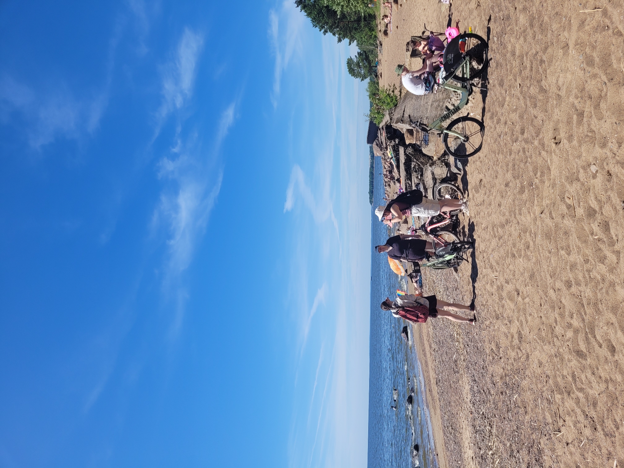 The image depicts a sunny day at a beach in Repino, Russia, with the Gulf of Finland in the background. Several people are present, engaged in various activities. In the foreground, two women stand near bicycles on the sandy beach. One woman is wearing a red backpack and a white top, while the other is wearing a dark shirt and a cap. Two bicycles are parked nearby. Further down the beach, a man and a child are seated in chairs. In the water, a few people are swimming. The beach is a mix of sand and pebbles, with some rocks and vegetation along the waterline. The sky is clear blue with scattered white clouds. The overall scene conveys a relaxed, recreational atmosphere on a summer day.