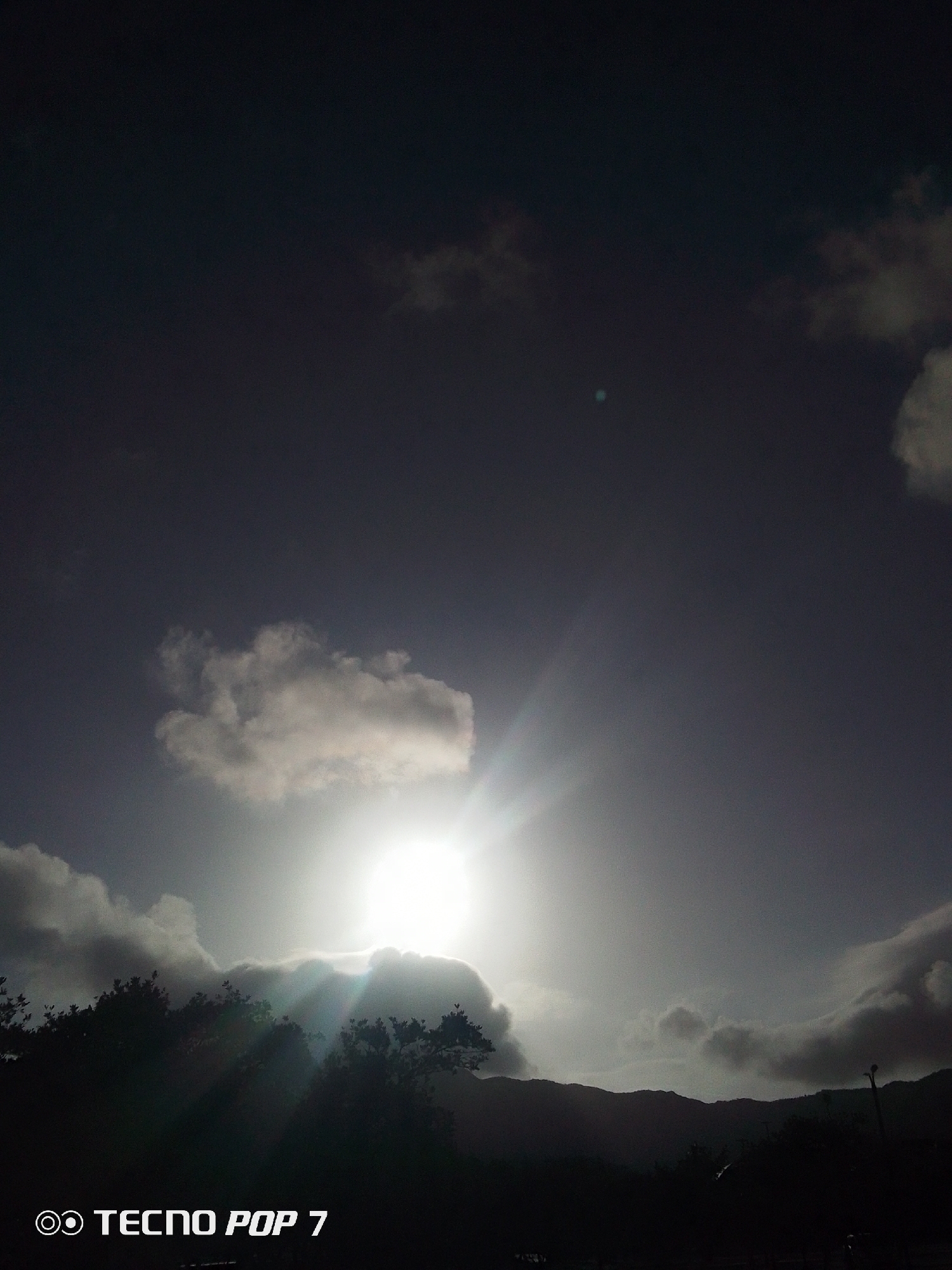 The image shows a sun obscured by clouds, taken during daylight in Pedro González, Venezuela. The sky is a mix of dark gray and light gray from the clouds. The sun's rays peek through the clouds. Silhouetted trees and mountains are in the foreground. A light pole is on the right of the mountain range. The words "Tecno Pop 7" are in the lower-left corner.