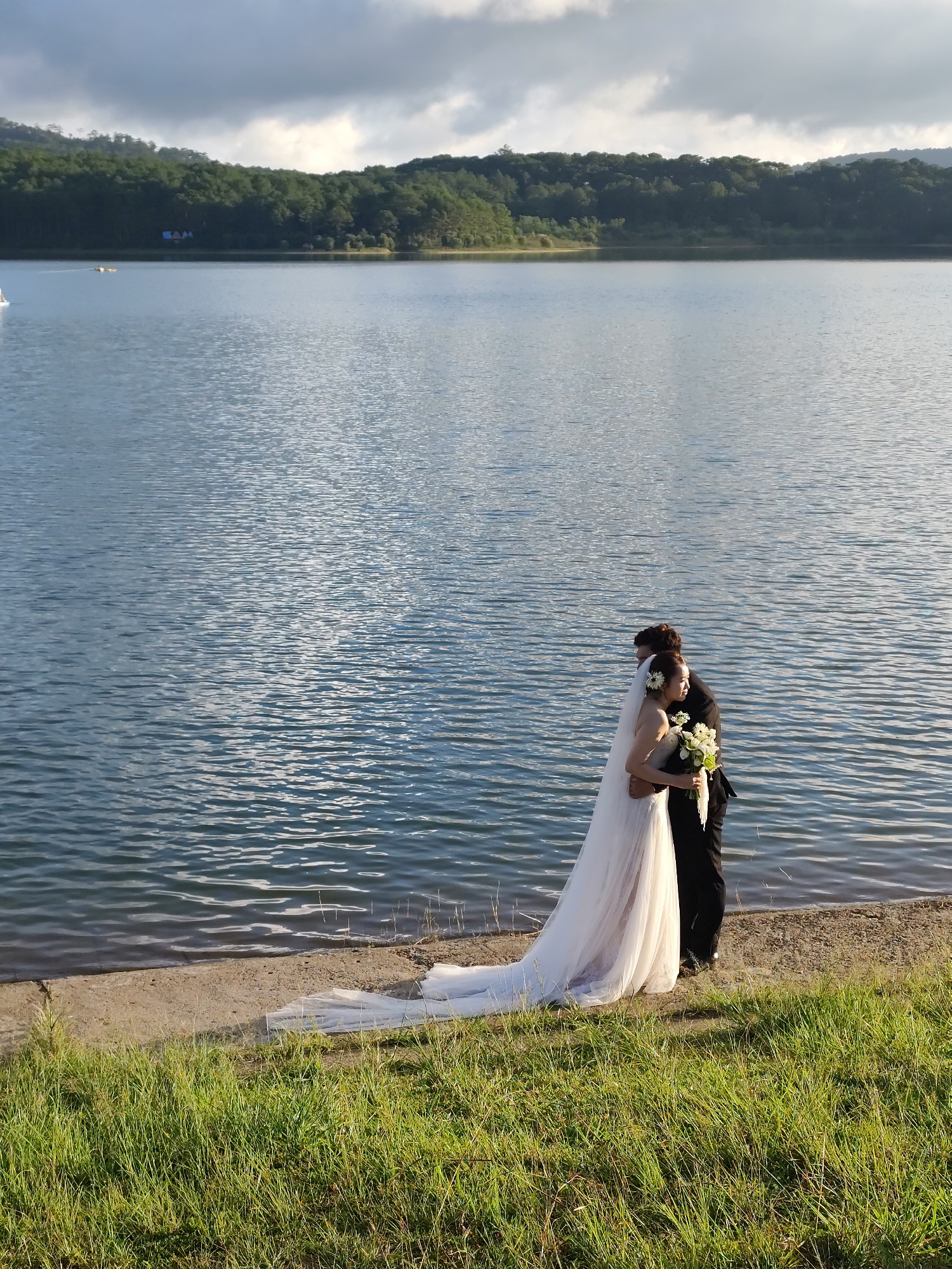A photograph captures a couple embracing on the shore of Tuyen Lam Lake in Da Lat, Vietnam. The bride, in a white wedding gown and veil, holds a bouquet of white and yellow flowers. The groom stands beside her in a black suit. The lake, occupying much of the background, is surrounded by green, forested hills. The foreground is characterized by a grassy embankment leading down to the sandy shoreline where the couple stands. Above, the sky is partly cloudy.