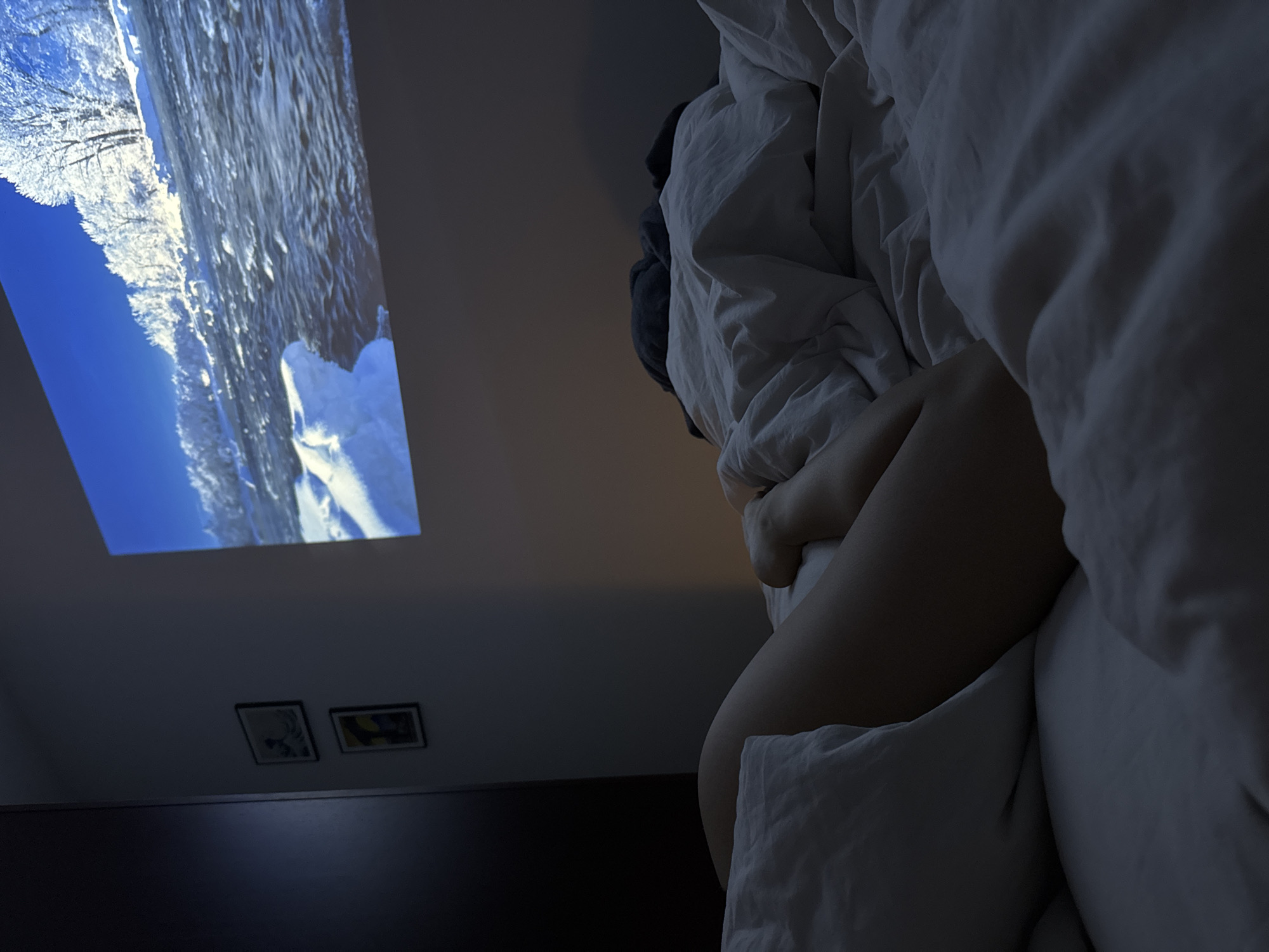 The image shows a person in bed in a darkened room, with a projection of a winter landscape displayed on the ceiling. The person is under a white duvet, with a portion of their leg visible. Two framed pictures hang on the wall below the projection. The room appears to be a bedroom with dark walls and minimal lighting. The location is not discernible from the image content alone and cannot be confirmed to be in Warsaw, Poland.