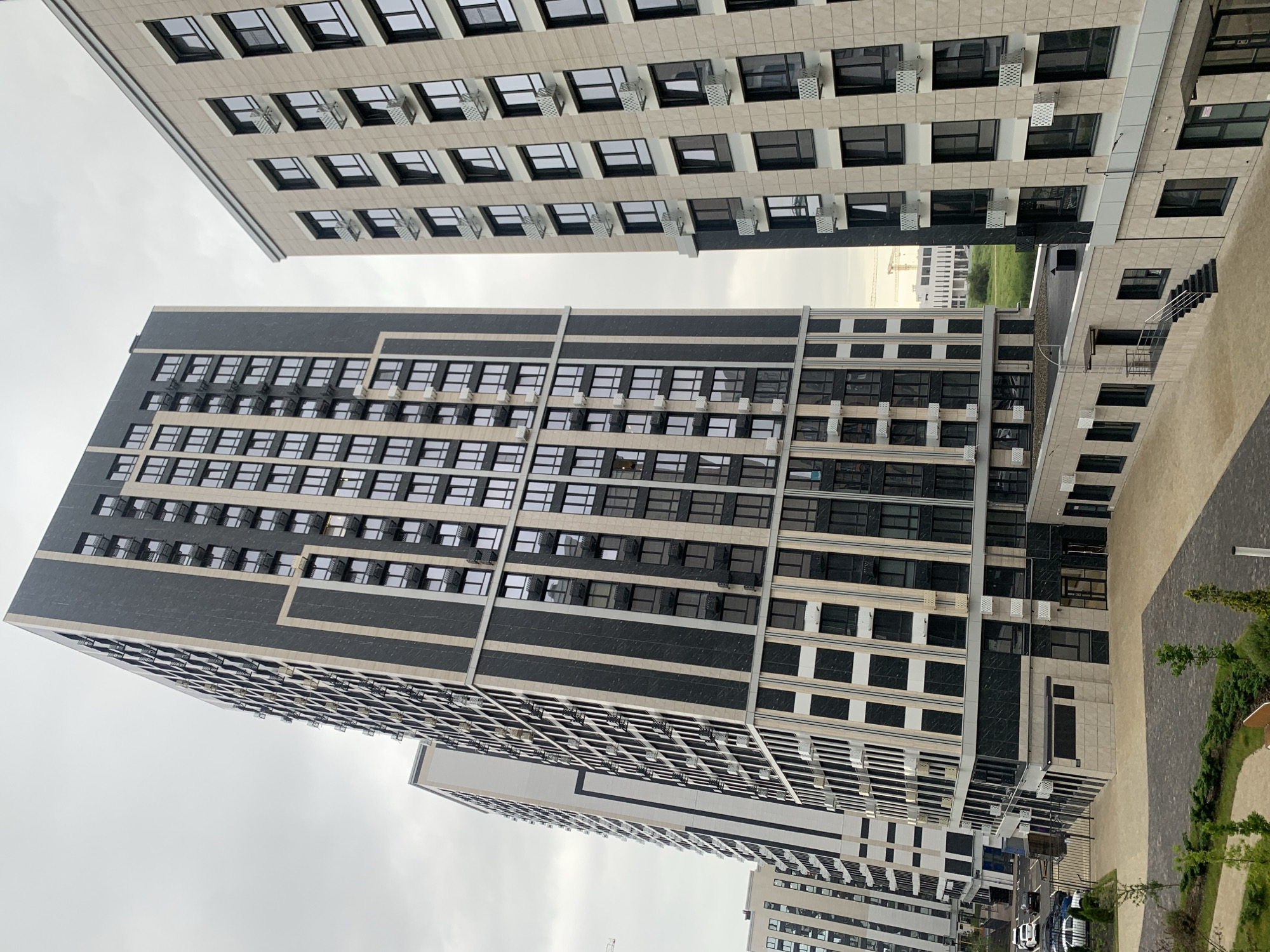 This media file showcases a modern urban landscape in Stavropol, Russia, dominated by residential buildings. The primary subjects are the imposing, multi-story apartment complexes with their repetitive facades of windows and balconies. The architecture features a blend of light-colored stone-like cladding and darker, possibly granite, sections, with metallic accents. Many windows are visible, suggesting a densely populated residential area.
The scene appears to be captured during daylight, under an overcast sky, indicated by the diffused lighting and lack of strong shadows. The weather seems to be cloudy or hazy. The setting is clearly an outdoor, developed urban environment, likely a courtyard or street between the buildings. The perspective is from a slightly elevated viewpoint, looking up at the structures and also down into some of the lower areas, revealing paved walkways, greenery, and parked cars.
Notable details include the numerous air conditioning units mounted on the exterior walls of the buildings, characteristic of modern apartment blocks. The arrangement of windows and balconies is highly geometric and uniform across the structures. In the background, glimpses of other buildings and construction cranes suggest ongoing development or a large, newly built residential district. The image is devoid of any visible people or specific events taking place, presenting a static view of the architecture and urban environment. There is no discernible text within the image. The overall impression is one of a contemporary, functional, and somewhat impersonal residential complex.