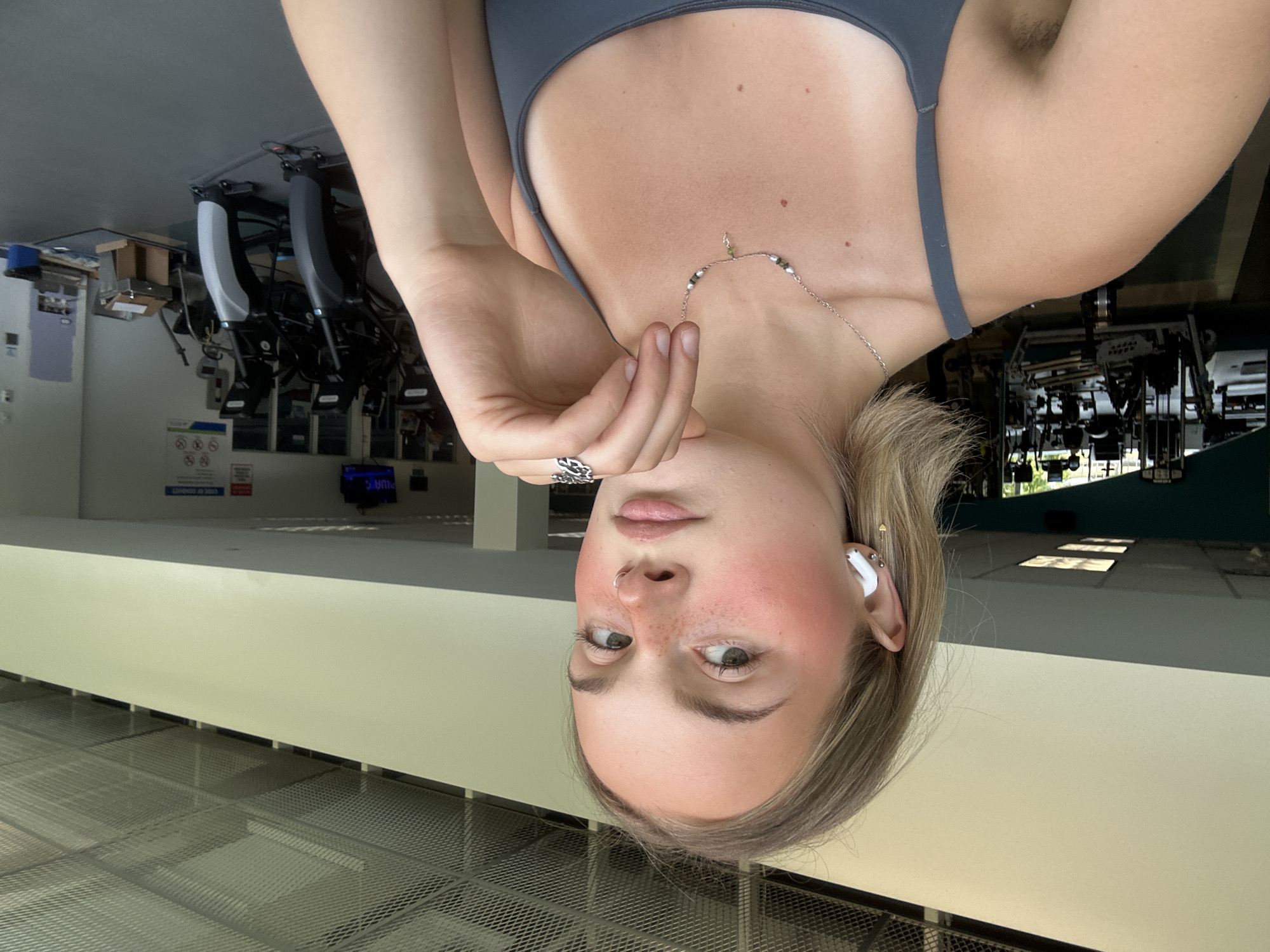 A young woman with blonde hair is taking a selfie in a gym. She is wearing a gray sports bra and has on white earbuds, a necklace, and rings. Her left hand is resting on her chin and her right hand is near her face. The gym has various exercise machines in the background. The location is not Whistler, Canada.