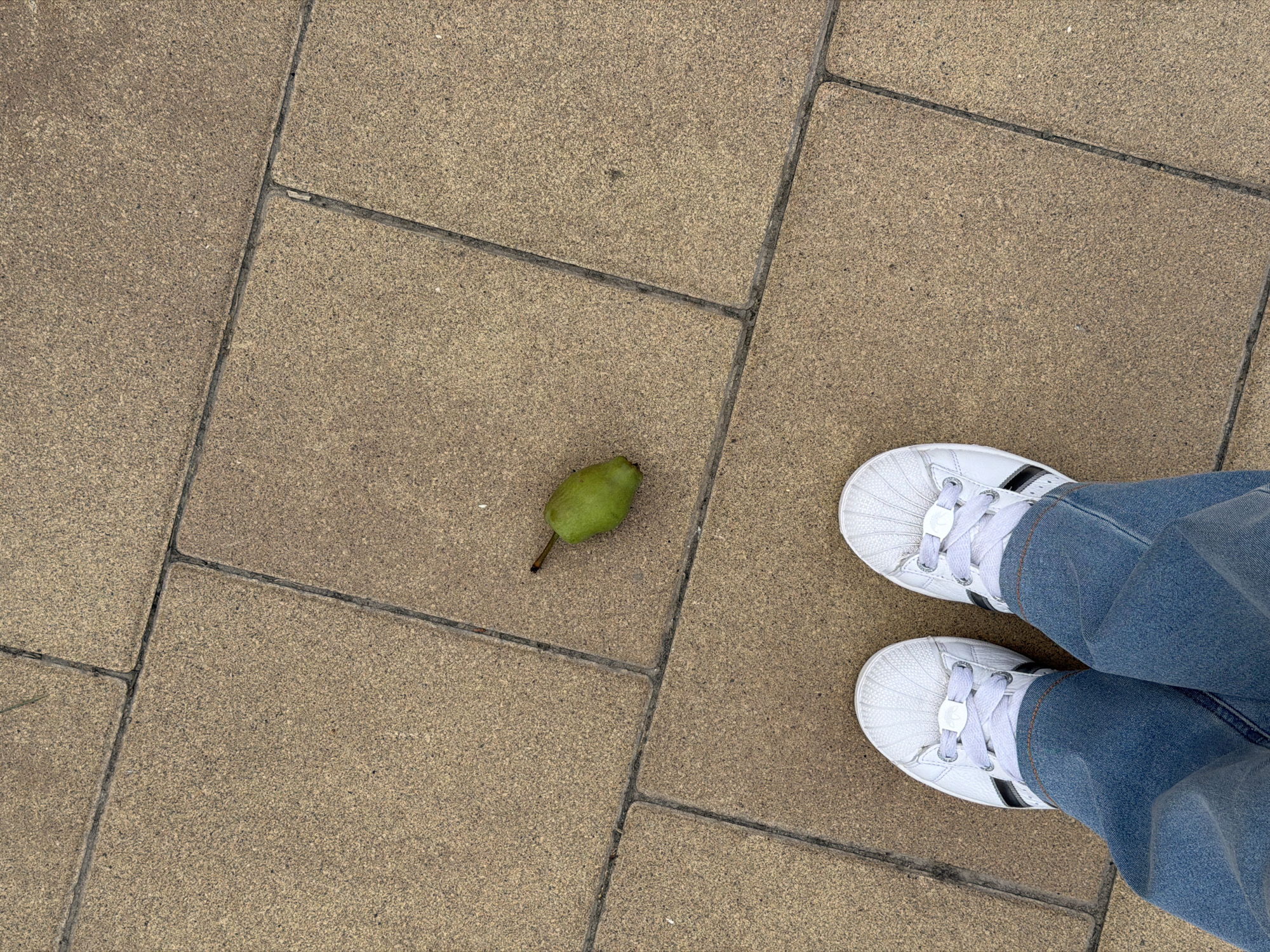 A medium shot captures a person's feet clad in white Adidas sneakers and blue jeans, standing on a paved ground. The ground is composed of rectangular tan pavers, laid in a grid pattern with dark grout lines. To the left of the shoes, a single, small, green pear lies on the pavers. The scene appears to be outdoors, likely on a sidewalk or pedestrian area. No specific landmarks identify the location as Kyiv, Ukraine.