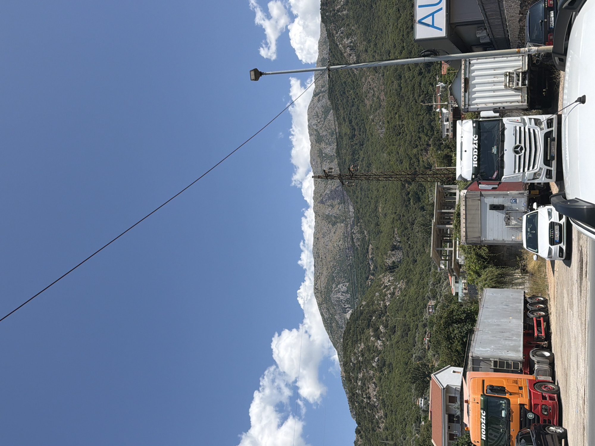 The image depicts a street scene in Zaljevo, Montenegro, under a clear blue sky with scattered white clouds. In the foreground, several large trucks are parked. The most prominent is an orange and red semi-trailer truck with "HODZIC" written on its cab. To its right, a white Mercedes-Benz semi-trailer truck, also with "HODZIC" branding, is parked. Behind these trucks, other trailers and vehicles are visible.
A white Audi sedan is parked in the middle ground, facing towards the right of the frame. Further back, residential buildings with red-tiled roofs are nestled into the side of a lush green mountain. In the background, the imposing, rocky face of the mountain dominates the landscape, its slopes covered in dense vegetation. A power line stretches diagonally across the upper portion of the image, and utility poles are also present. A street lamp stands to the right of the frame. The overall impression is one of a busy service area or transportation hub situated at the base of a scenic mountain range.