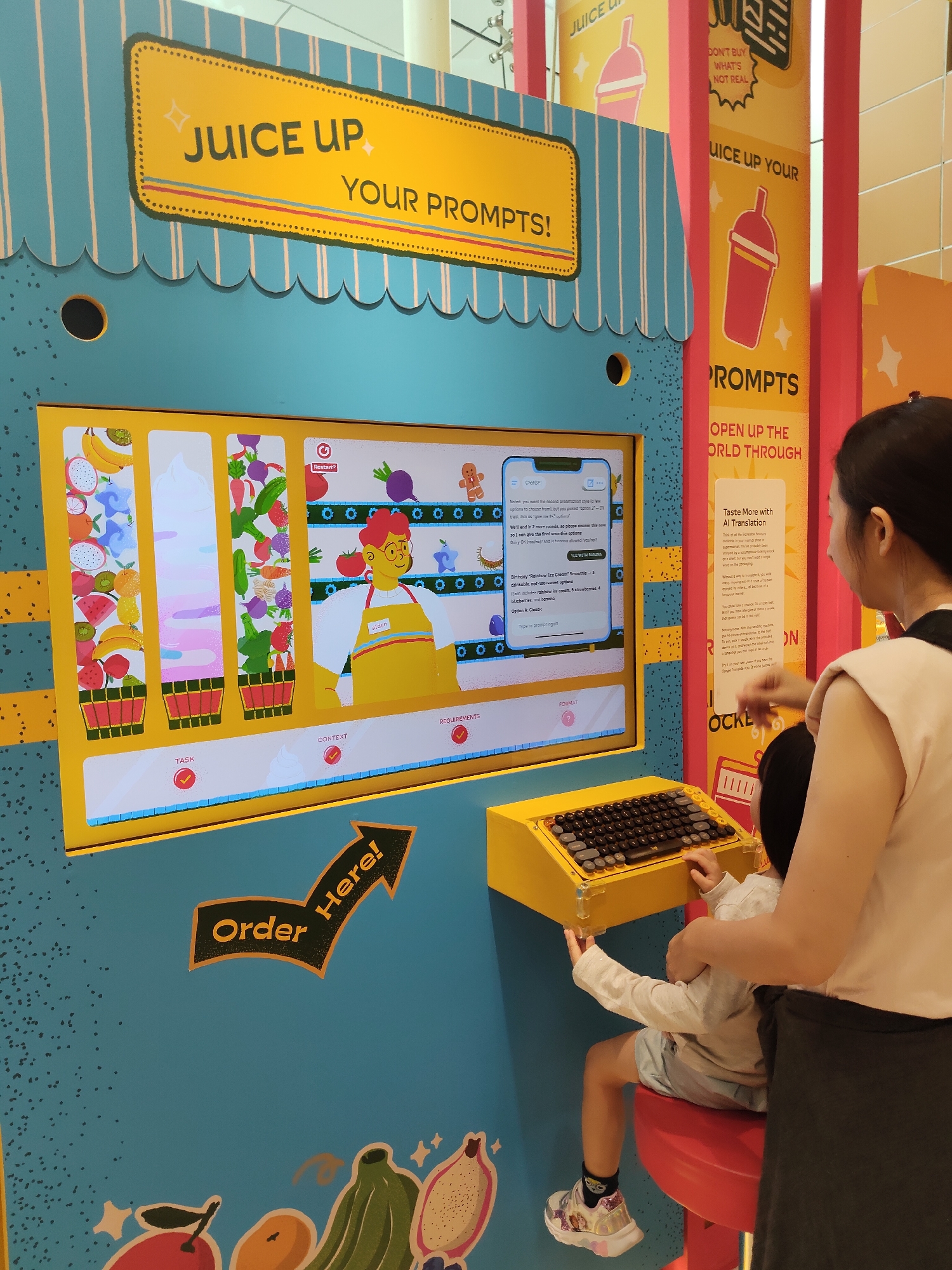 This is a photograph of an interactive exhibit in Singapore. The exhibit, themed "Juice Up Your Prompts!", features a large, colorful screen displaying a digital juice-making interface. On the screen, a virtual character named "Aiden" wearing an apron stands beside rows of animated fruits and vegetables. To the right of the screen, a woman and a child are interacting with the exhibit. The child is seated on a red stool, operating a yellow keyboard. The woman stands behind the child, guiding their interaction with the keyboard. The exhibit is designed to be engaging and educational, likely teaching users about prompt engineering or AI translation through a playful, gamified experience. The overall environment appears to be a public space, possibly a museum or a family-friendly attraction.