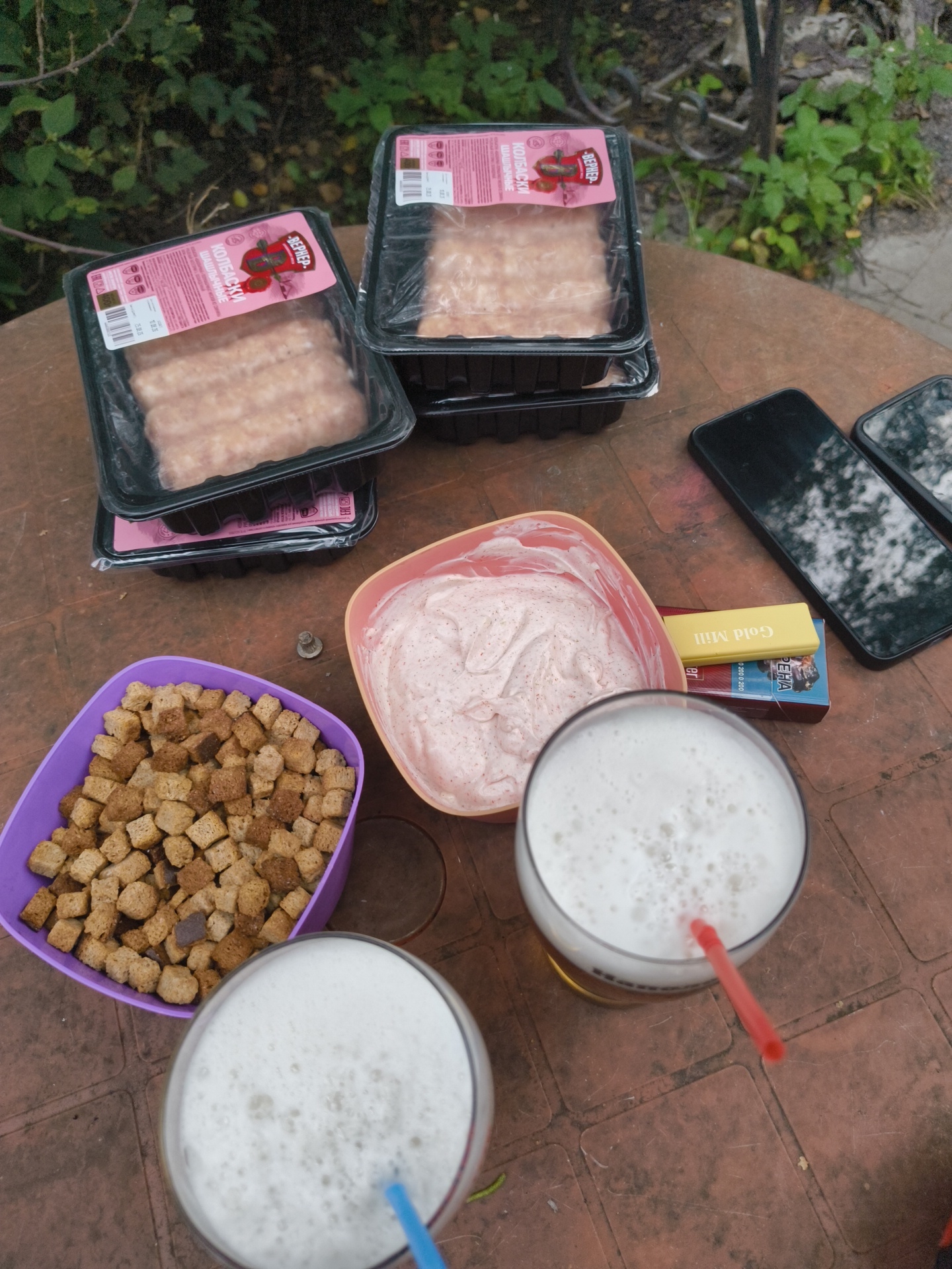 Two glasses of frothy, light-colored beverage, likely beer, are positioned on a textured, brown, outdoor table. One glass is in the foreground, with a red straw submerged in its foamy head. Behind it, another glass with a blue straw is visible. To the left of the glasses, a purple plastic container is filled with small, cubed croutons. To the right of the croutons, a pinkish bowl contains a creamy dip with visible flecks. Further to the right, a yellow cigarette pack and a pack of cigarettes are placed on the table. Two dark-colored smartphones are also on the table, one partially obscuring the other. In the background, two packages of raw, pinkish sausage links are stacked on the table. The surroundings are overgrown with green foliage, suggesting an outdoor setting, possibly a yard or garden. The overall scene depicts an informal outdoor gathering with food and drinks.