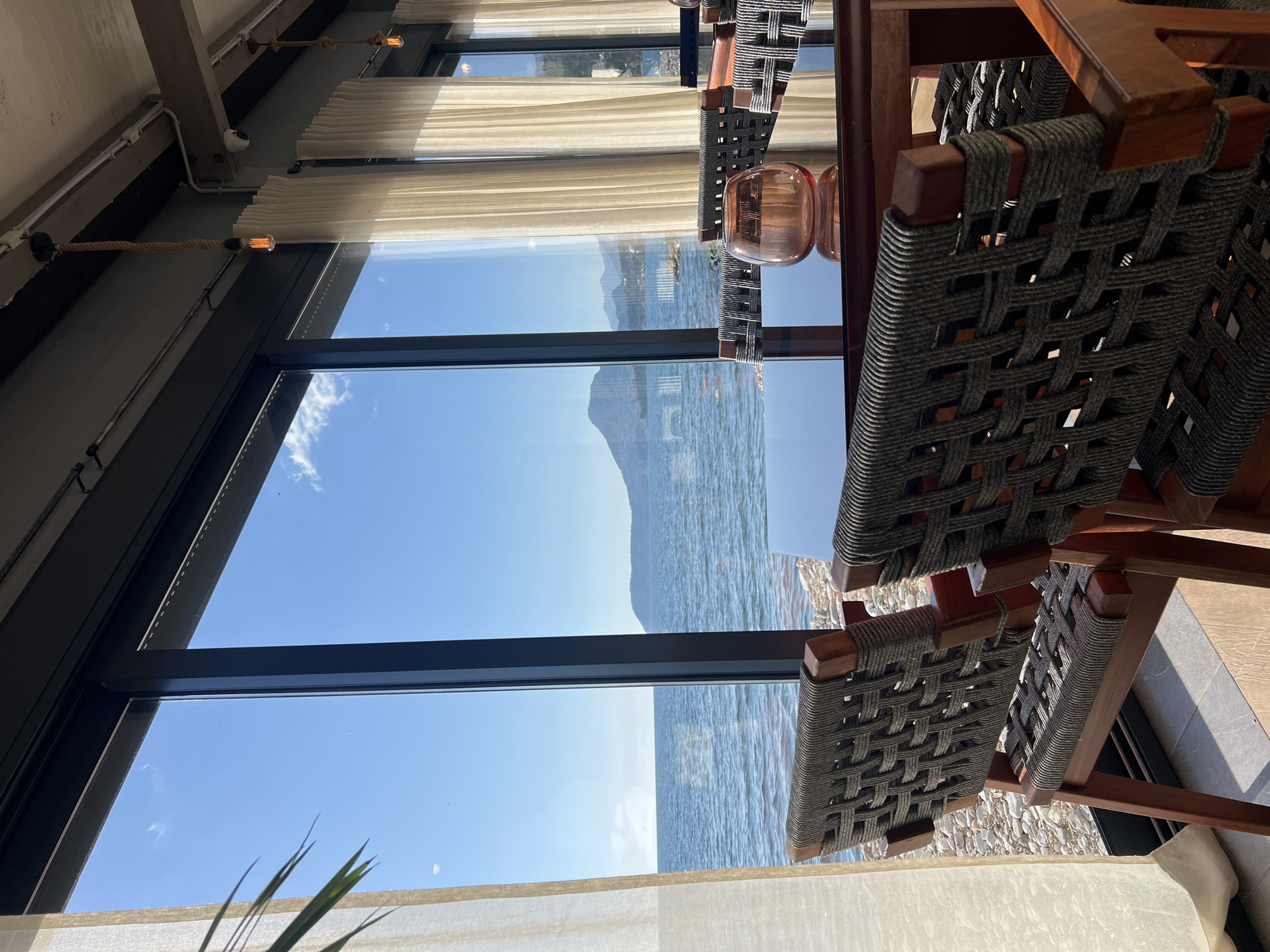 The image shows a view from what appears to be an indoor seating area in a location overlooking the Adriatic Sea in Bar, Montenegro. The primary focus is the view through a large window, where a calm sea meets a distant mountain or coastal landmass under a clear blue sky. Foreground elements include woven-seat chairs and a glass table, partially obscuring the view. Light-colored curtains adorn the window, and there are hints of interior architectural details like ceiling fixtures. The overall scene creates an impression of an elegant dining or lounge space with scenic views.