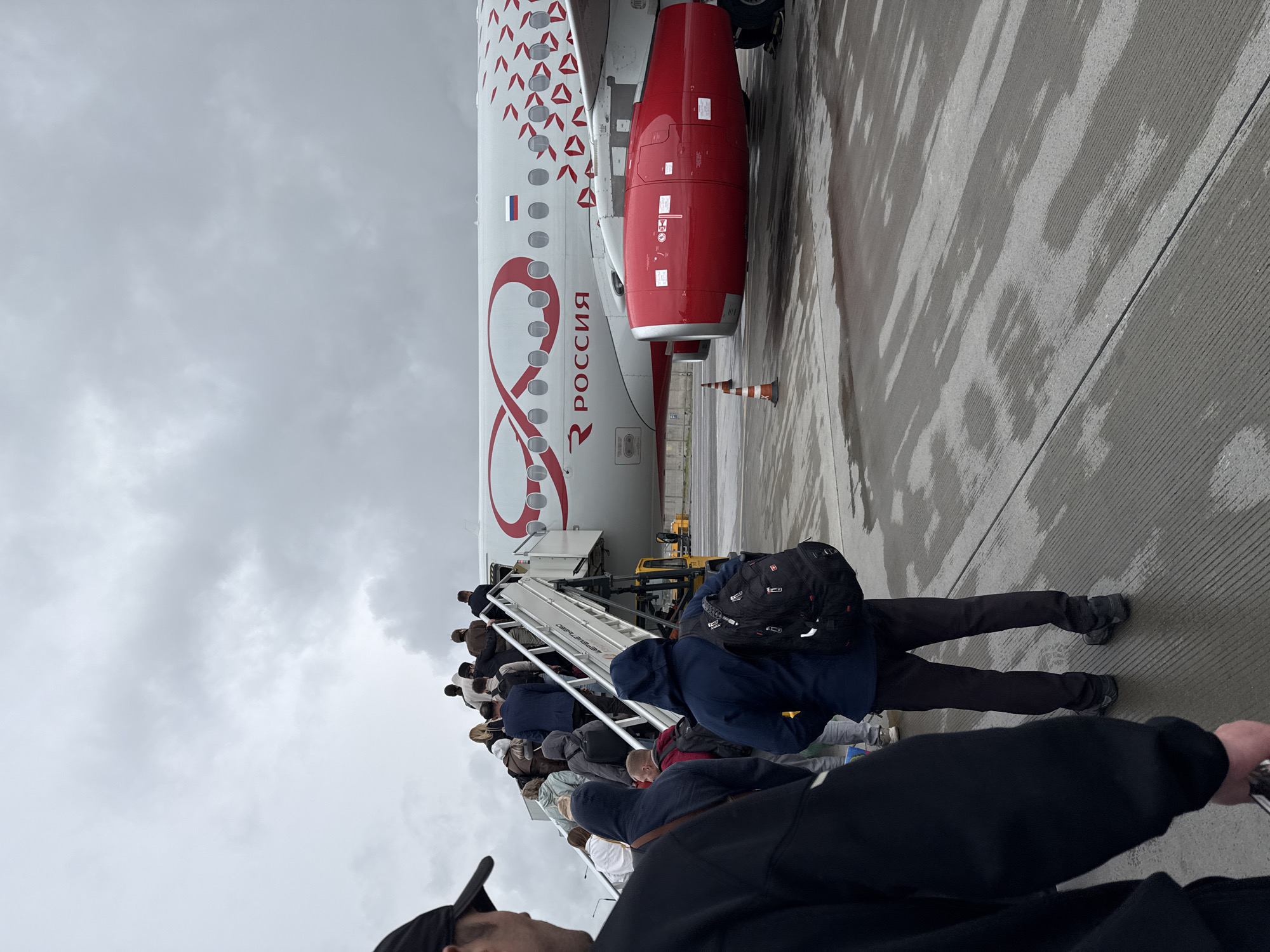 The image shows passengers boarding a Rossiya Airlines airplane via mobile stairs. The plane's tail section, a red engine, and the "Россия" inscription on its side are visible. People are lined up on the stairs to board the plane. The sky is overcast. The location is likely the Sheremetyevo International Airport in Khimki, Russia, based on Rossiya Airlines operating from there.