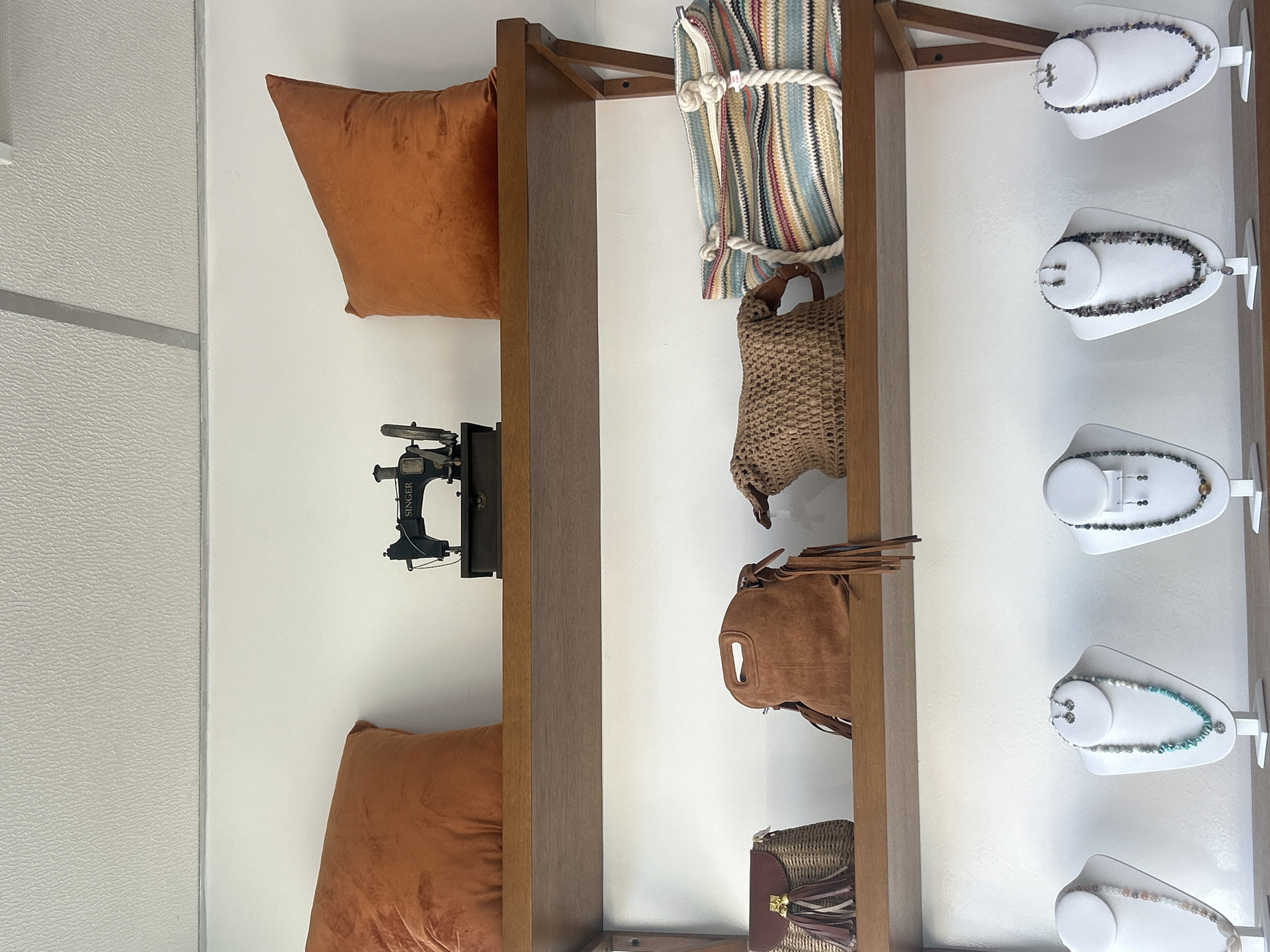 The image depicts a vertically oriented display in an interior setting. On the left side, there is a white wall with a textured surface, adjacent to a wooden shelving unit. The shelving unit houses an orange pillow, a black vintage Singer sewing machine, a striped tote bag secured with rope, a woven handbag, a brown leather backpack with fringe, and a wicker purse with leather accents. To the right of the shelving unit, necklaces on white bust stands are displayed against another white wall. The location and city are unspecified.