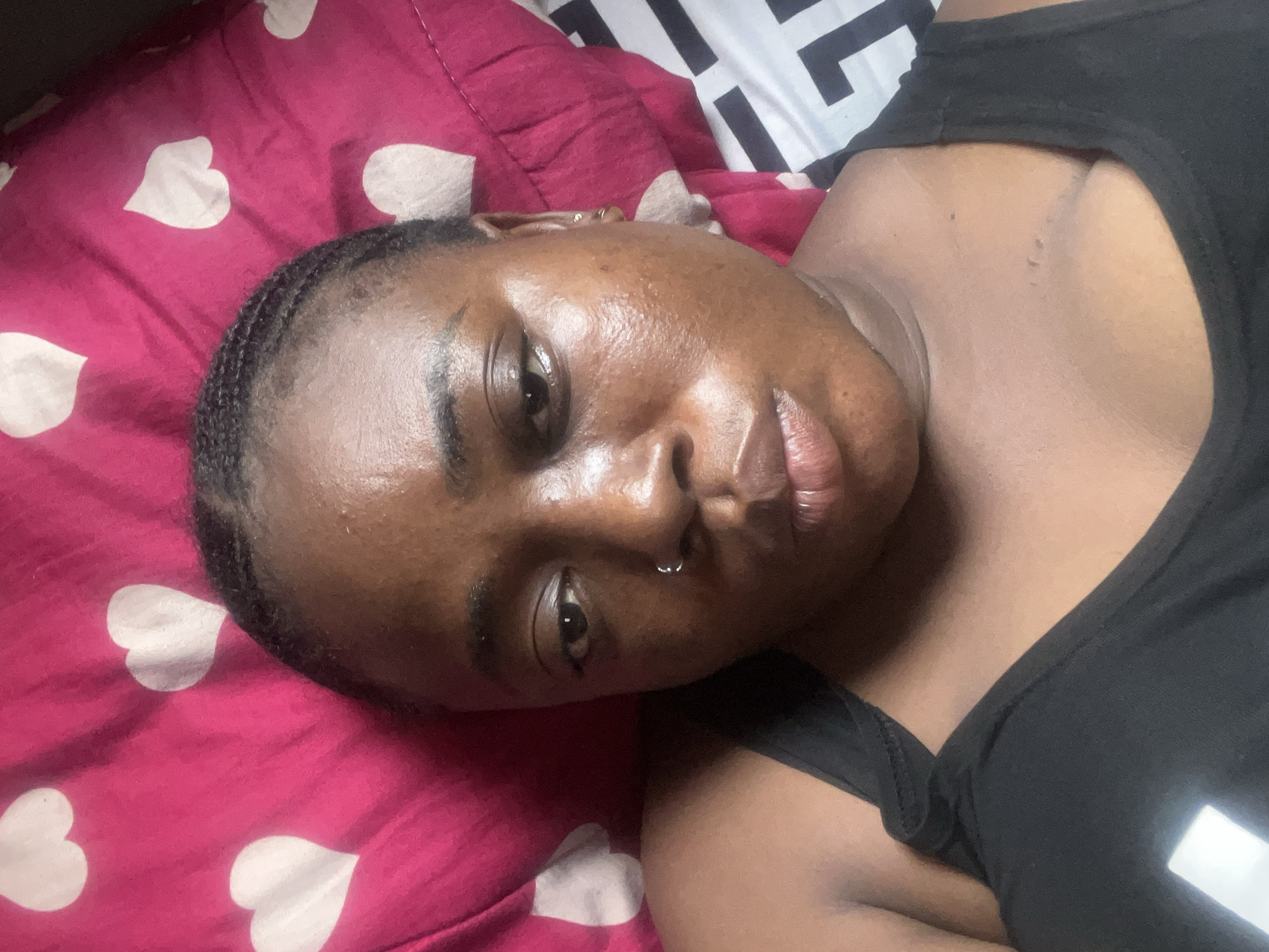 A close-up shot shows a Black woman lying on a bed. She wears a black tank top and has short, braided hair. A silver ring is visible in her left nostril. Her skin has a shiny, oily appearance. The bed has two different pillowcases. The one closer to her head is magenta with white heart shapes. The other has a black and white geometric design. There is no indication of this photo being taken in Abuja, Nigeria.