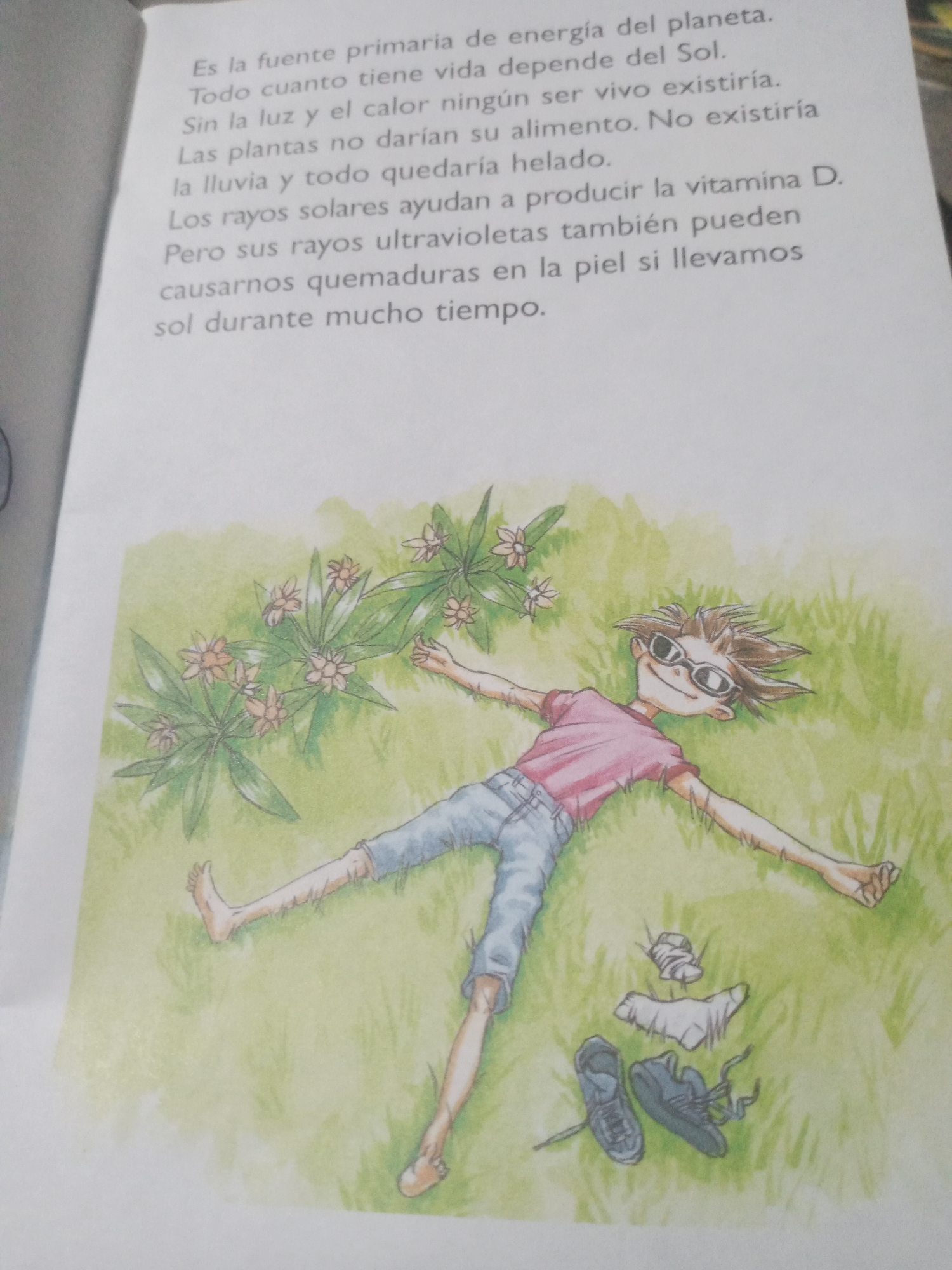 The image displays an open book with text on the left page and an illustration on the right page. The text, written in Spanish, discusses the sun as the primary source of energy for the planet and the dependence of life on it, as well as the role of sun rays in producing vitamin D and their potential to cause sunburn. The illustration depicts a young person lying on their back in a grassy field, with arms and legs spread out. The person is wearing a pink t-shirt, blue jeans, and sunglasses. Scattered near the person's feet are a pair of dark sneakers and some crumpled white socks. In the background, a small plant with pink flowers is visible. The scene is rendered in a colorful, cartoonish style. There is no information within the image or its context that indicates a specific location in the city of Santa Ana, Venezuela.