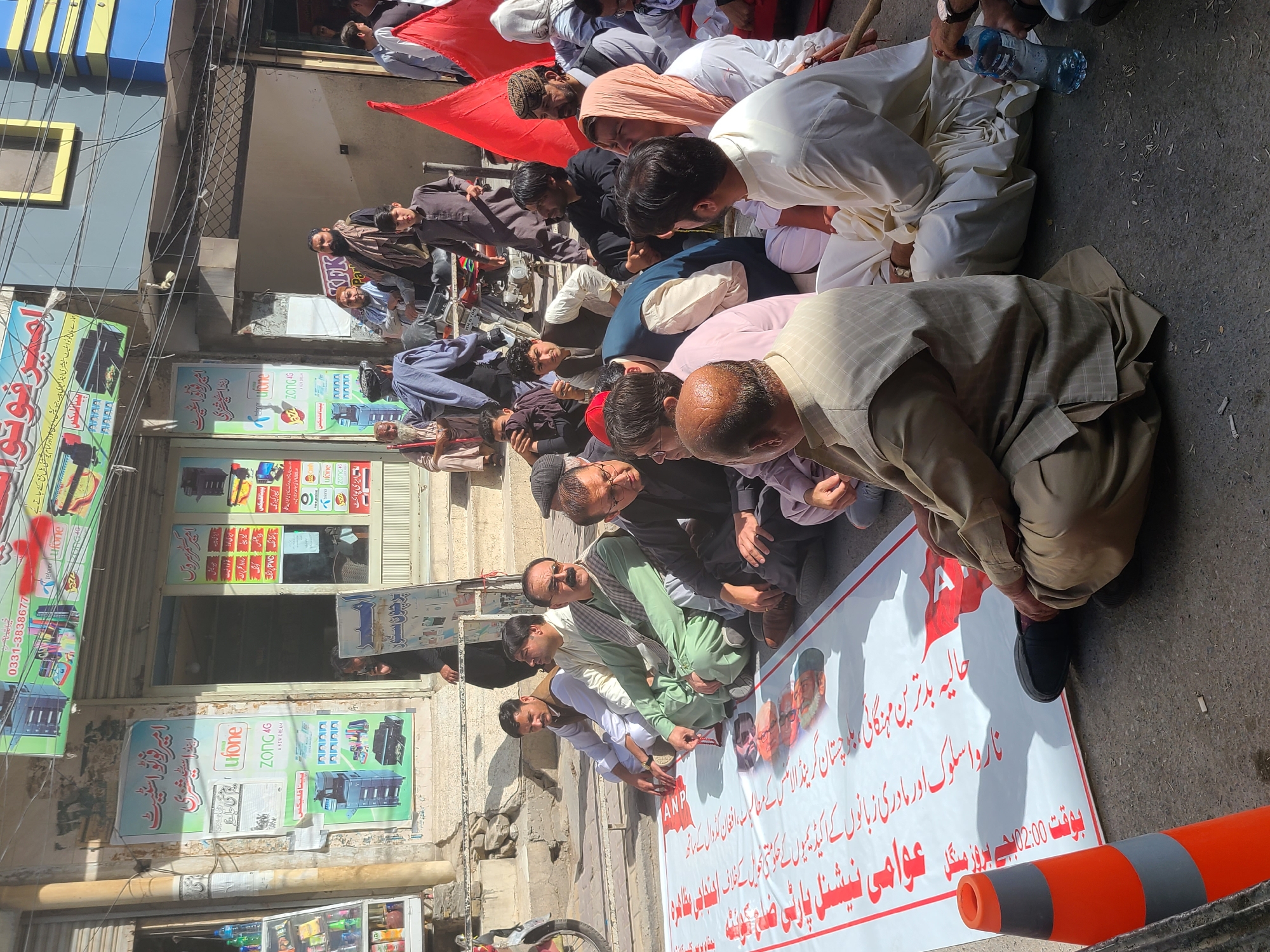 A group of men are sitting or kneeling on a street in Quetta City Tehsil, Pakistan, around a large white banner with red lettering and images of people. The banner appears to be associated with the Awami National Party. Several of the men are interacting with the banner, possibly signing it. A red flag is visible behind them. The surroundings suggest an urban commercial area, with storefronts and utility lines overhead. Some of the storefronts have signs for businesses such as "Ufone" and "Zong 4G."