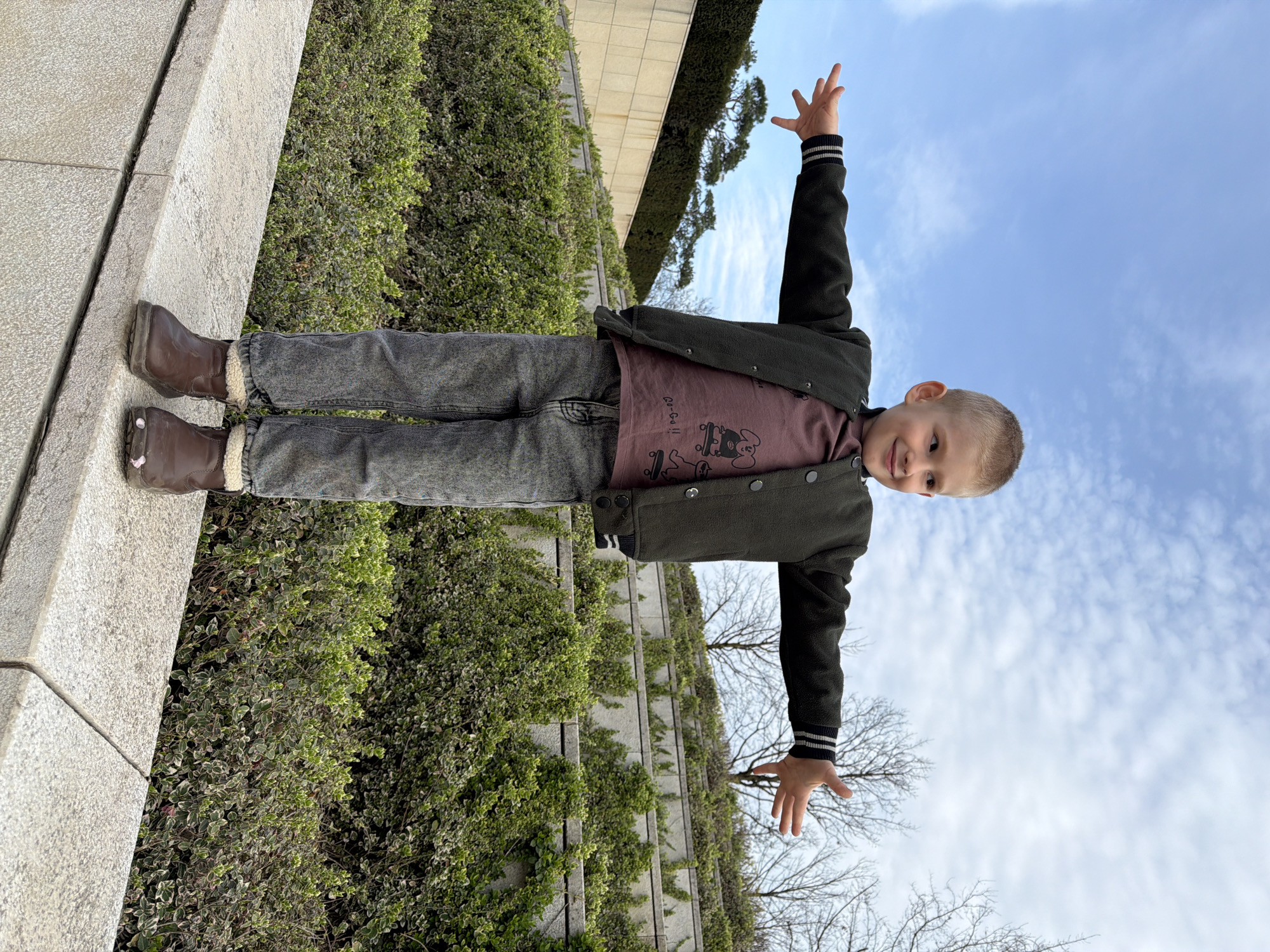 A boy is standing on a ledge in an outdoor setting, possibly in Krasnodar, Russia. He is wearing a dark jacket, a maroon t-shirt with a graphic design, gray jeans, and brown boots. His arms are extended outwards and upwards. The background consists of green vegetation, concrete structures, and a blue sky with scattered clouds. The boy appears to be posing or playing.