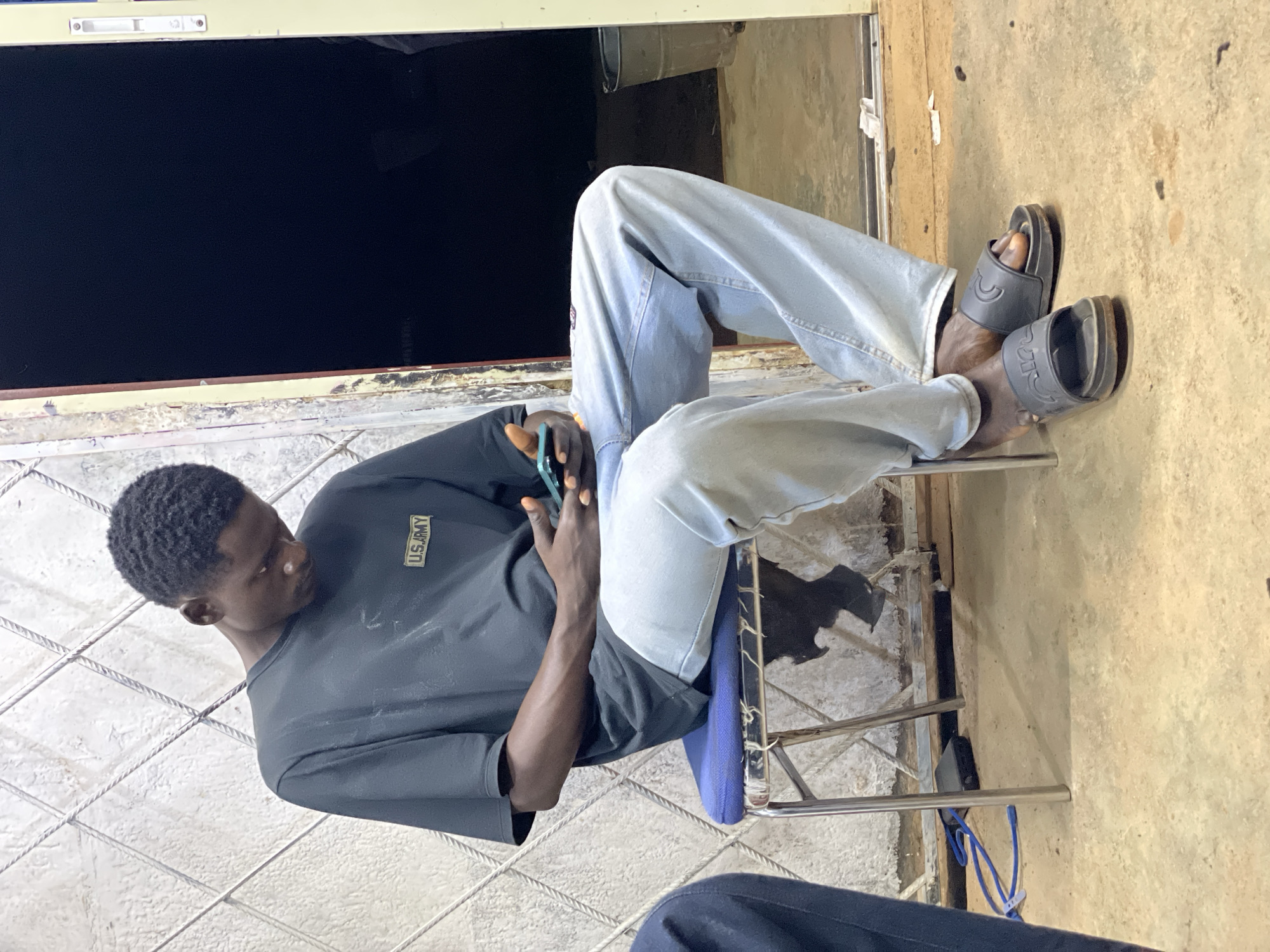 A young Black male wearing a dark t-shirt with a U.S. ARMY patch, light blue jeans, and black slides sits on a blue-cushioned metal stool. He is looking down at a smartphone in his hands. The background consists of a textured white wall with a diamond pattern of wire, a doorway revealing darkness, and a concrete floor. A blue cord and a black rectangular object are on the floor near the stool. The scene appears to be outdoors, possibly in a utilitarian or informal setting within Durumi, Nigeria.