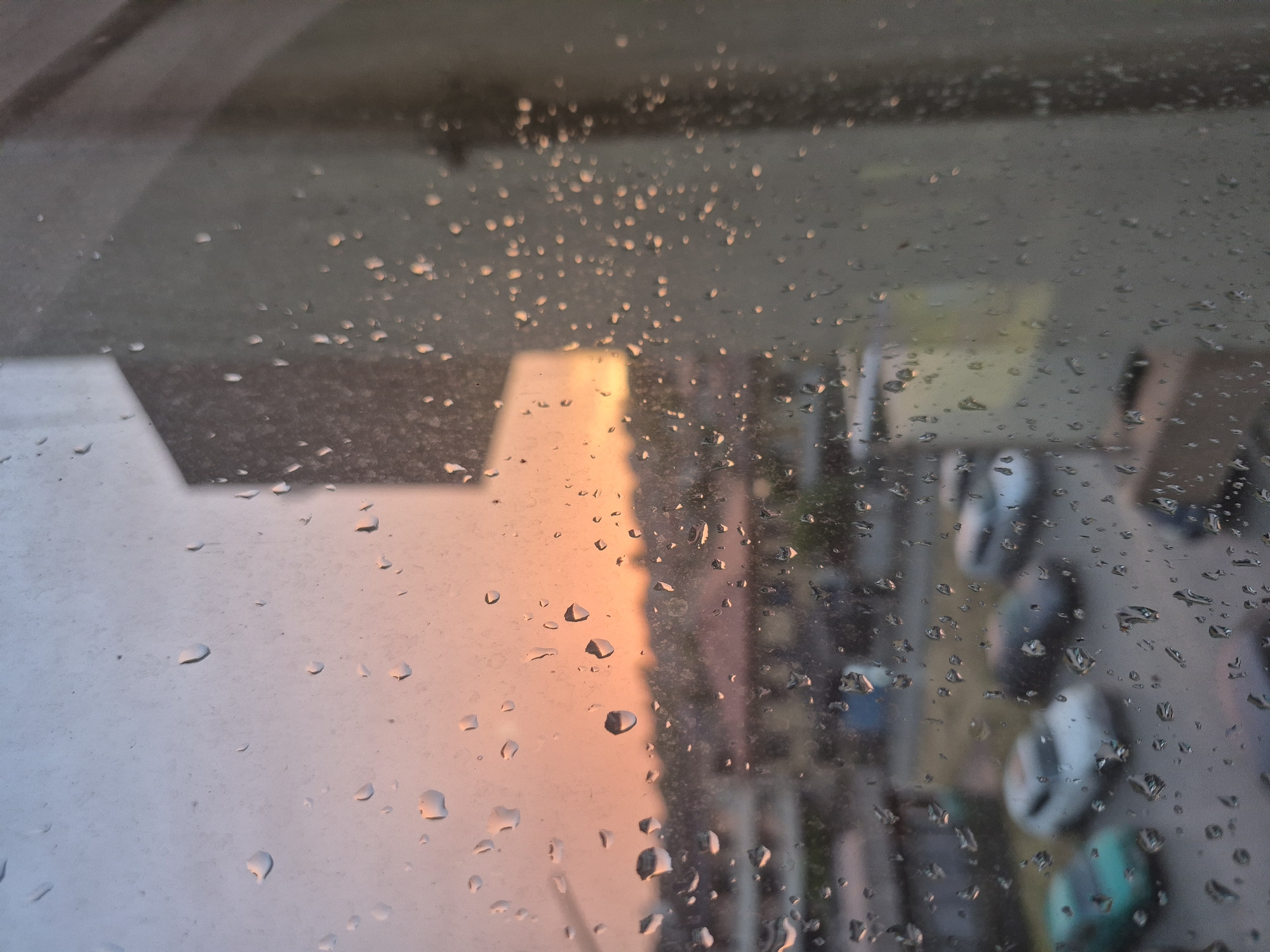 The image captures a view through a rain-streaked window, overlooking a street scene in Popovo, Bulgaria. The time of day appears to be dusk, with the sky displaying soft hues of orange and pink. In the foreground, water droplets of varying sizes are scattered across the glass, distorting the view of the environment beyond.
The middle ground reveals a street with several parked cars, including a teal-colored vehicle on the left, a white car next to it, and darker vehicles further down the street. The buildings in the background are low-rise structures, typical of a residential area in a city like Popovo. The overall impression is one of a quiet, possibly post-rain, evening in the Bulgarian town. There are no people visible in the frame, and no specific actions or interactions are taking place. The environment is urban, characterized by buildings and vehicles, with the atmospheric condition of rain evident on the window.