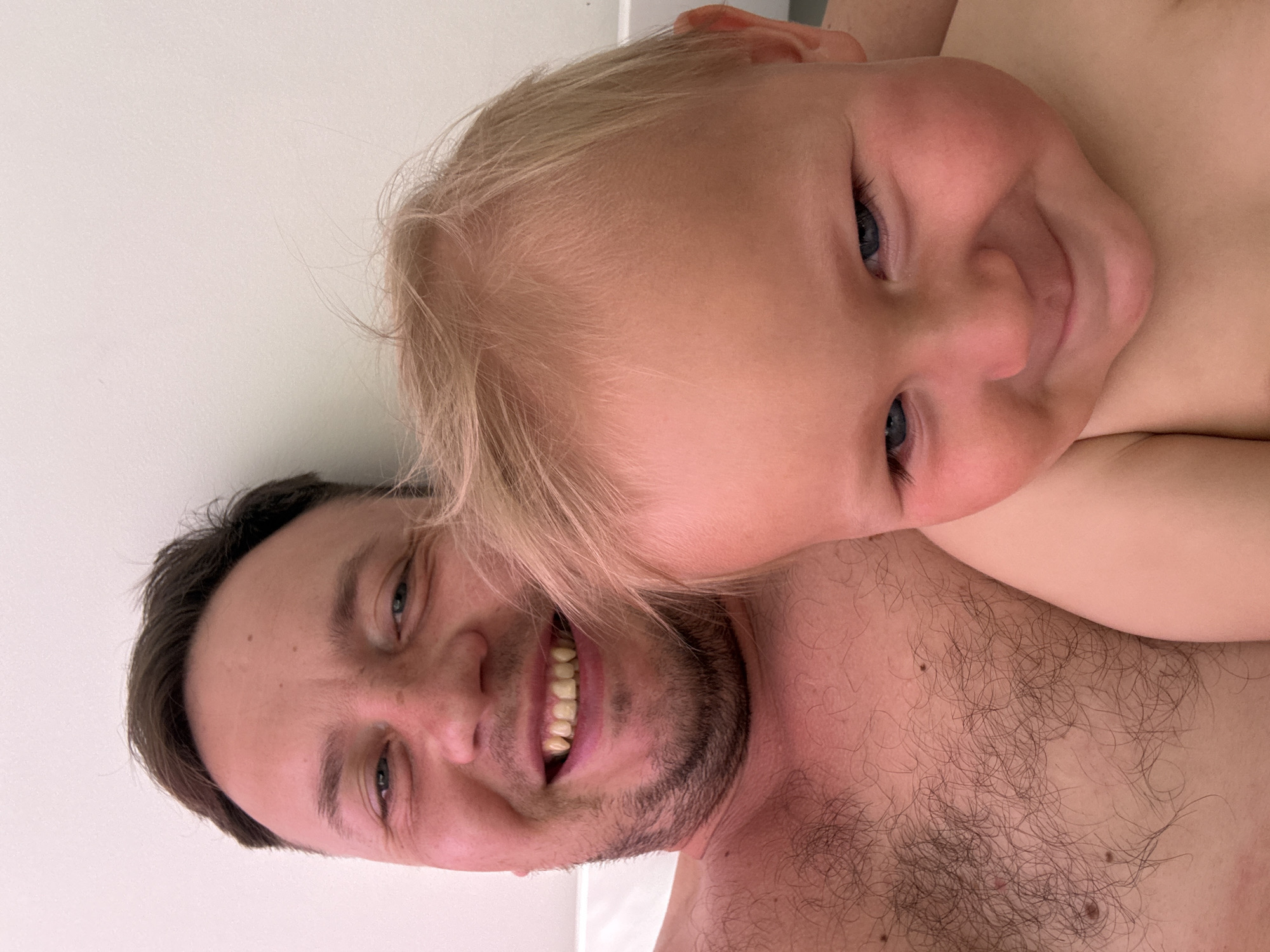 In a close-up selfie, a man and a young child are seen with their faces filling most of the frame against a plain, light-colored background. The man, positioned on the left, smiles broadly, revealing his teeth, and has dark hair and stubble. His bare chest, covered in dark hair, is visible. The child, on the right, has blonde hair and blue eyes, and is also smiling. The image appears to be taken indoors, possibly in a home, with the clean background suggesting a simple environment. There are no identifiable landmarks or specific details that would indicate a location within Sochi, Russia. The overall scene captures a moment of shared joy and intimacy between the two individuals.