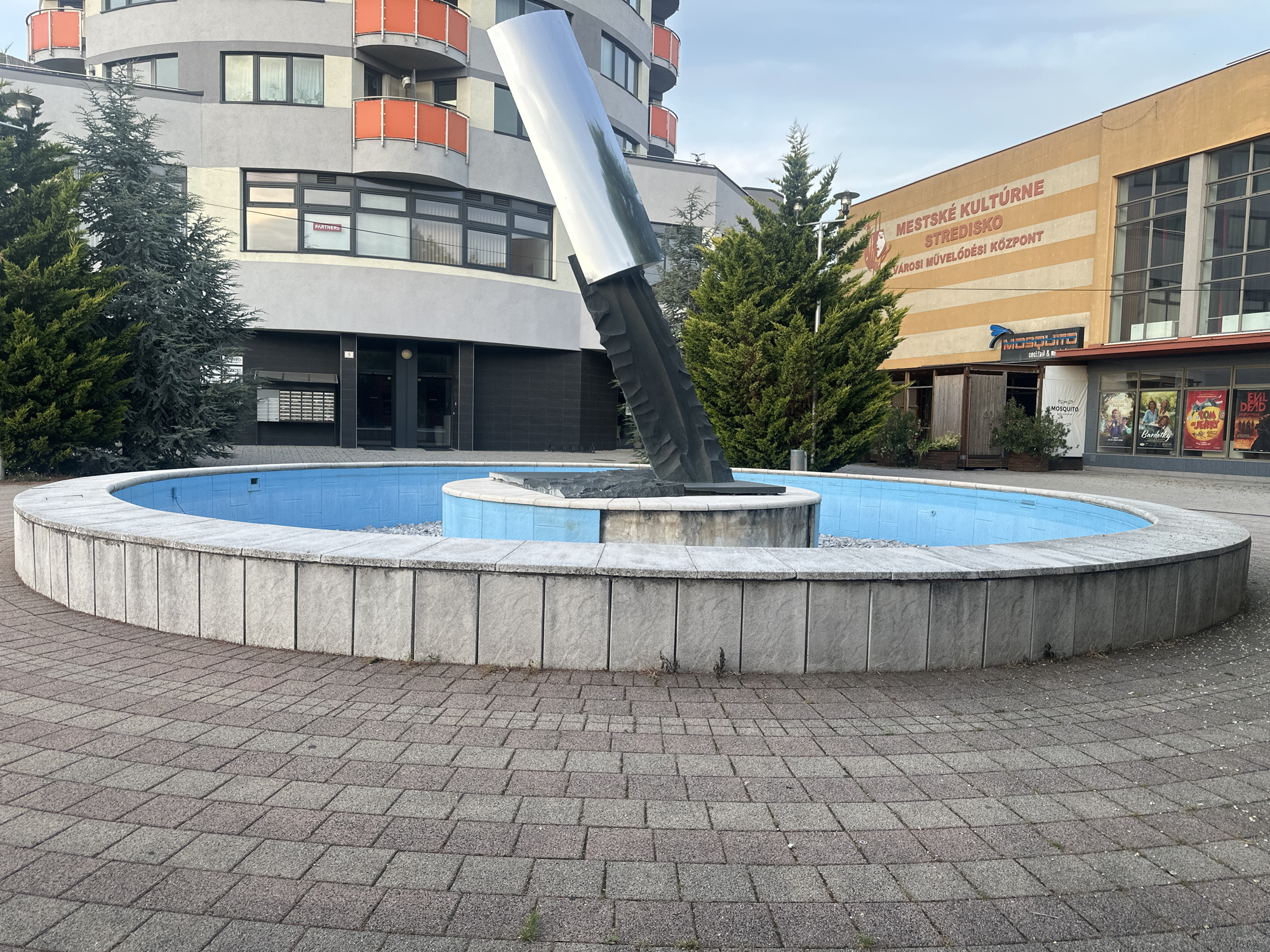 An outdoor, ground-level, wide shot captures a circular fountain in the foreground, with a modern apartment building and a cultural center behind it. The fountain, filled with blue water, is surrounded by a low, light gray stone wall. The area around the fountain is paved with interlocking rectangular bricks in shades of gray and reddish-brown.
Dominating the center of the fountain is a tall, abstract metal sculpture. The sculpture has a dark, rough-textured base that appears to be carved or shaped, from which a highly polished, reflective silver piece extends diagonally upwards. The silver section reflects the sky and surrounding trees.
To the left of the fountain is a multi-story apartment building with a curved facade. It is painted in shades of gray, with orange balconies and balconies visible on several levels. Dark framed windows are scattered across the building. To the right of the apartment building, and behind the fountain, is a light yellow building with orange stripes. This building has large windows and signage that reads "MESTSKÉ KULTÚRNE STREDISKO" and "VÁROSI MŰVELŐDÉSI KÖZPONT". Below this, a blue and black sign for "MOSQUITO cocktail & more" is visible, and further to the right, movie posters are displayed.
Several evergreen trees, including tall, dark green spruces and shorter, lighter green cypress-like trees, are positioned around the fountain and between the buildings. The sky is overcast, suggesting it might be early morning or late afternoon. The overall scene depicts a public space in an urban setting, featuring modern architecture and a prominent art installation.