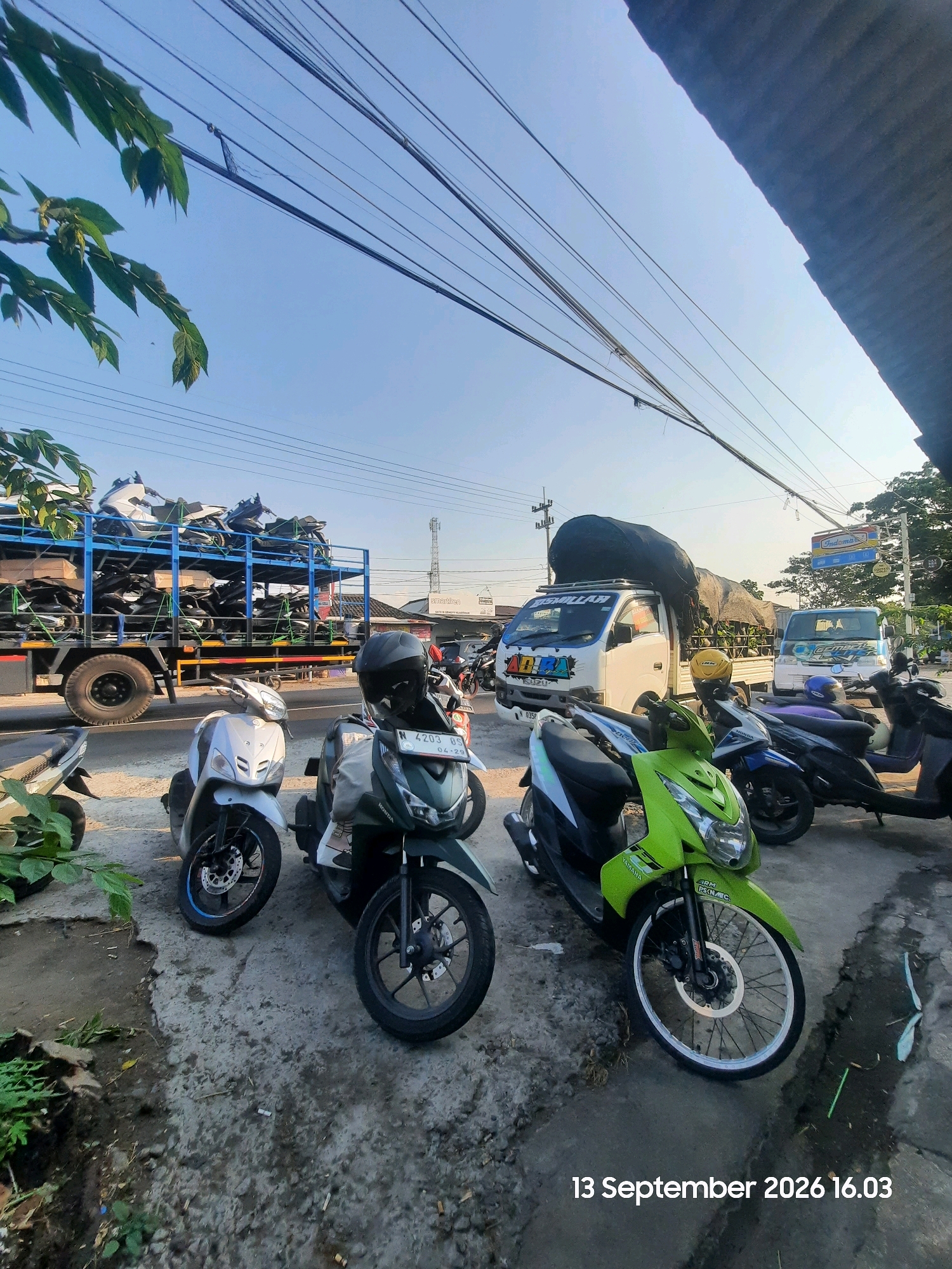 The image shows a street scene in Sukokerto, Indonesia, on September 13, 2026, at 4:03 PM. In the foreground, several parked motorbikes are visible, including a green Yamaha, a white scooter, and a dark-colored scooter. The motorbikes are parked on a paved area, with some vegetation and debris on the ground.
In the middle ground, a white Isuzu truck is parked, carrying a large, dark tarp-covered load. The truck has decals on its side, including the word "ALLAH" and what appears to be "ADABA". Another light blue truck with the word "CEMBONG" is parked behind it. In the background, a truck loaded with motorcycles on racks is parked along the road. The sky is clear and blue. The scene appears to be a roadside, possibly near a commercial area given the presence of the trucks and the Indomaret sign in the distance.
