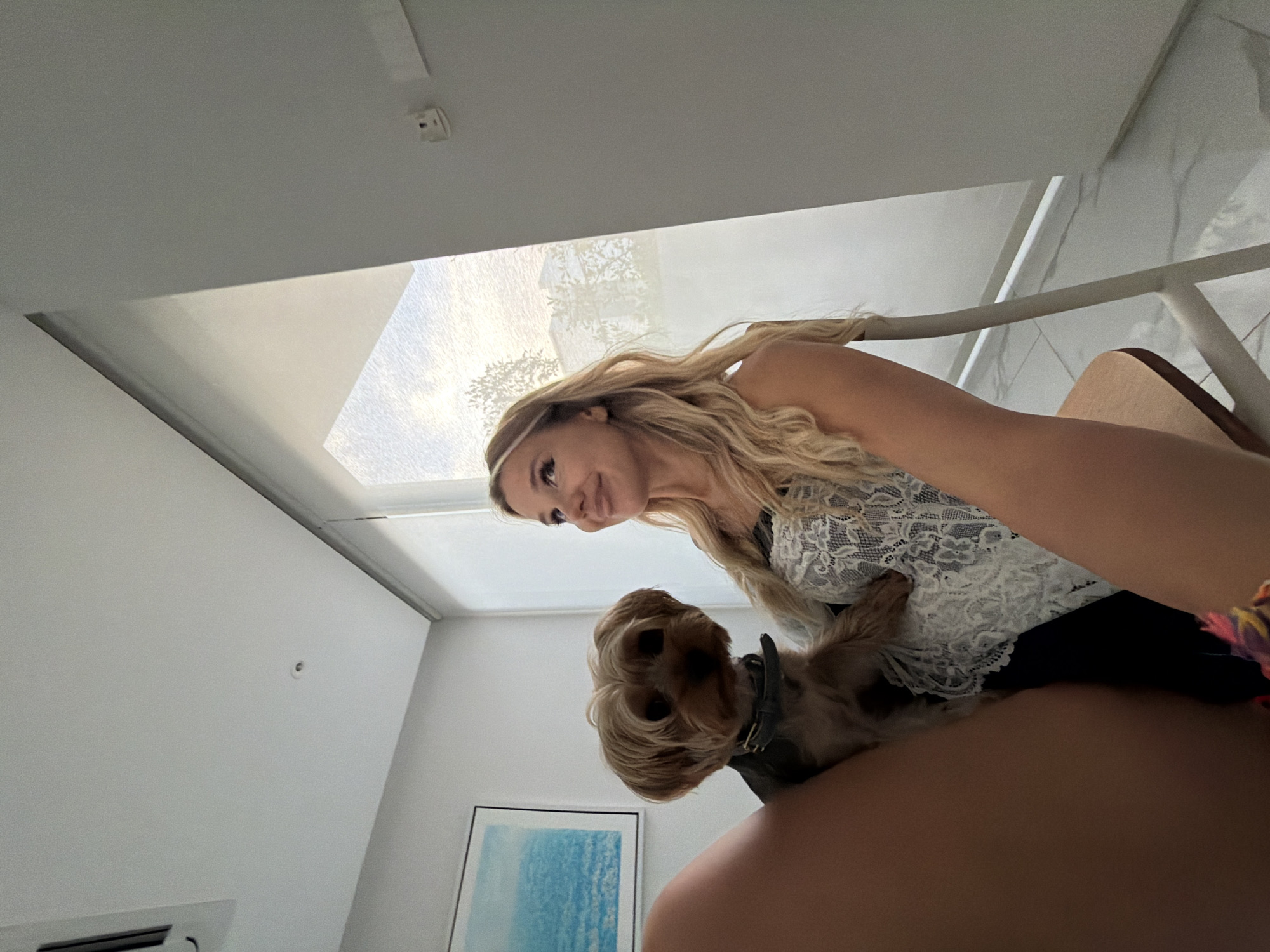 A woman with long blonde hair, wearing a white lace top, sits in a chair with a small Yorkshire Terrier in her lap. She is looking upwards and to the right, with a slight smile. The dog is looking forward. They are in an indoor setting with a large window with a sheer curtain, letting in natural light. A framed picture with blue tones hangs on the wall to the left. A white air conditioning unit is visible on the ceiling. The floor is tiled in a marble pattern. The overall atmosphere is casual and relaxed. The location is identified as Thep Krasatti, Thailand.