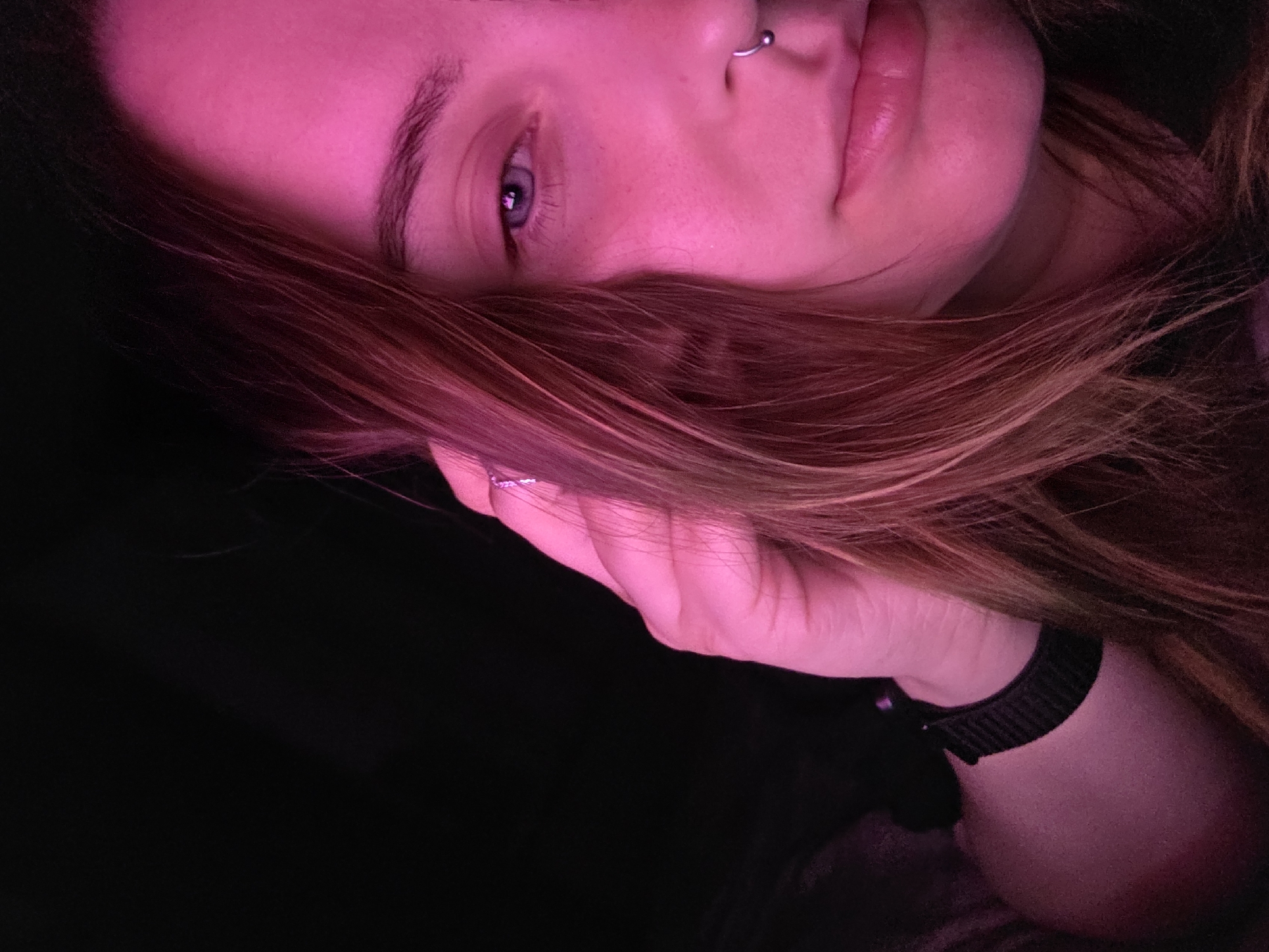 The image shows a close-up, angled view of a person's face illuminated by a pink light. The person has light skin, light brown or reddish-brown hair, and blue eyes. The person is wearing a nose ring. The person's hand is raised, with fingers visible. The person is wearing a dark-colored watch or wristband. The background is dark and out of focus. The location is Unknown, Unknown.