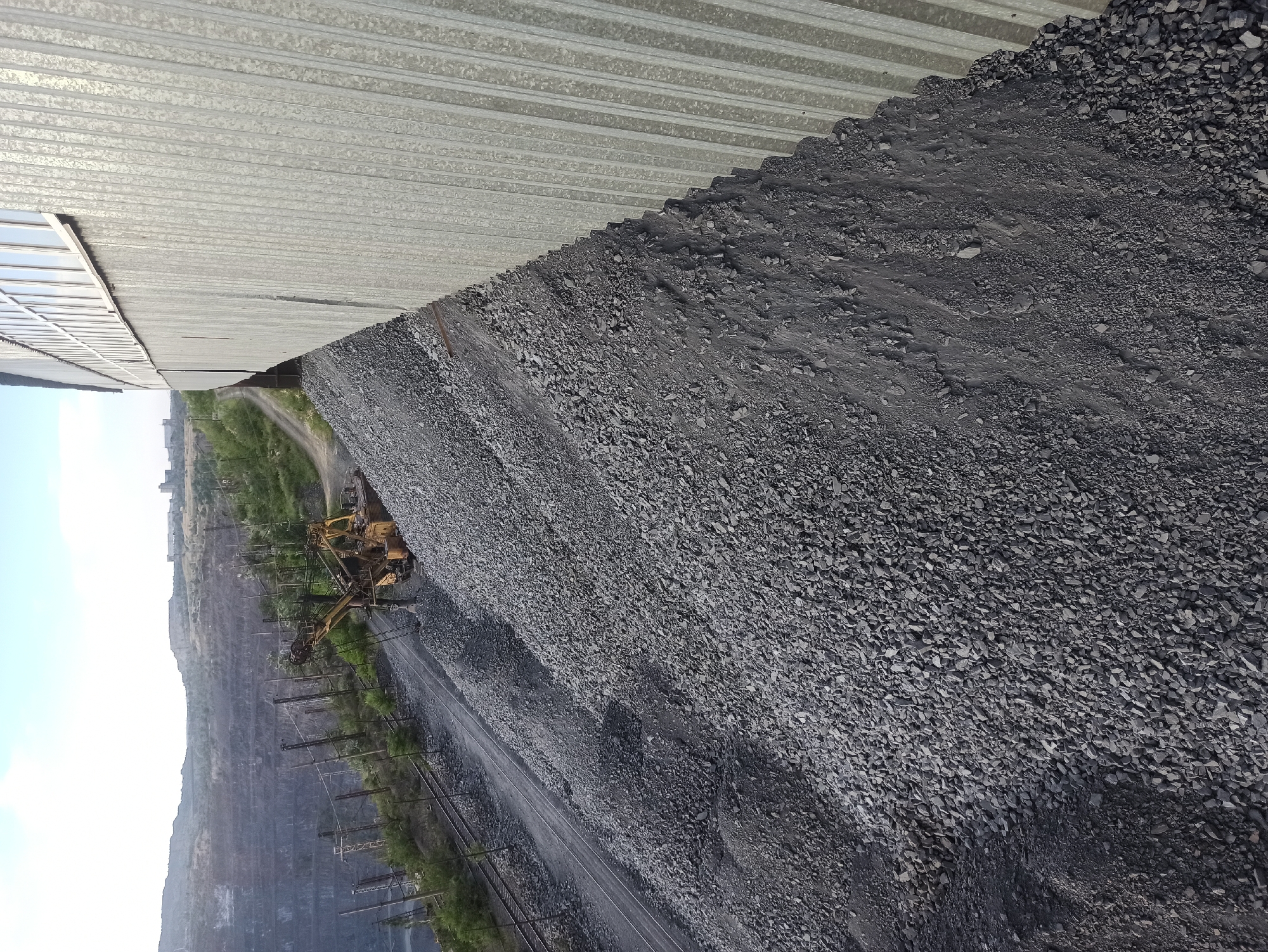 The image depicts an outdoor industrial scene, likely a quarry or mining operation, with a large pile of dark grey gravel dominating the foreground and mid-ground. The gravel is unevenly distributed, forming a steep slope that leads towards the background. To the right of the gravel pile, a corrugated metal wall of a building runs diagonally across the frame, extending from the bottom right corner towards the top right. The wall has a vertical ribbed texture and a row of windows along its upper edge.
In the mid-ground, to the left of the gravel pile, a yellow excavator is positioned on a railway track. The excavator appears to be stationary and partially covered in gravel. More railway tracks run parallel to the excavator and extend into the background. Beyond the tracks, a vast open-pit mine is visible, characterized by steep, layered walls and a hazy atmosphere. Distant structures and hills can be seen on the horizon. The sky above is a pale blue with some wispy clouds. The overall impression is one of a large-scale industrial operation with heavy machinery and vast quantities of excavated material.