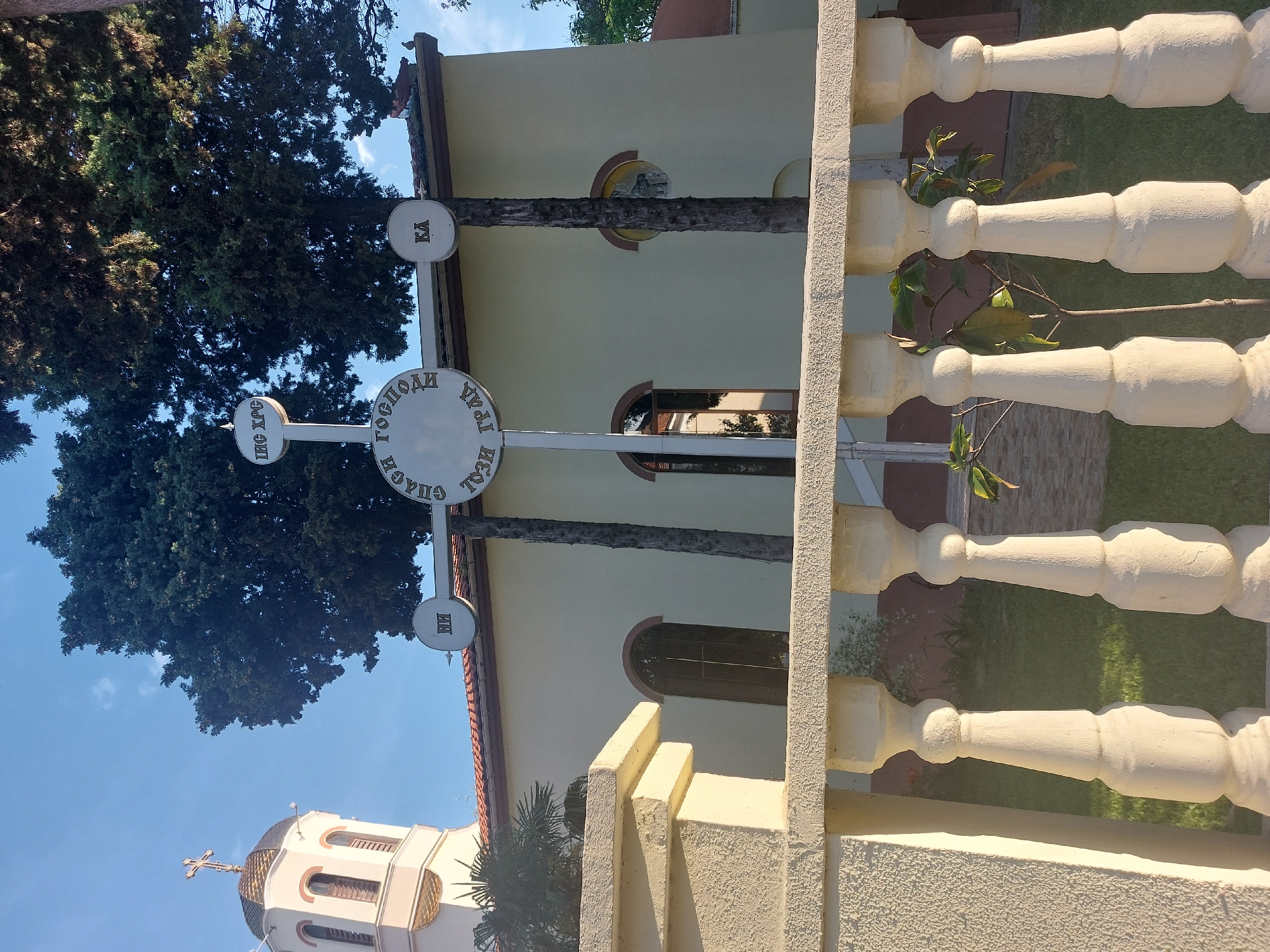 The image shows a view of the Church of the Holy Trinity in Tsarevo, Bulgaria. A large Orthodox cross with circular inscriptions in Cyrillic stands in front of the church. A tall tree partially obscures the view of the church building. A balustrade fence is in the foreground, slightly out of focus. The sky is blue and mostly clear. A small section of the church's bell tower is visible.