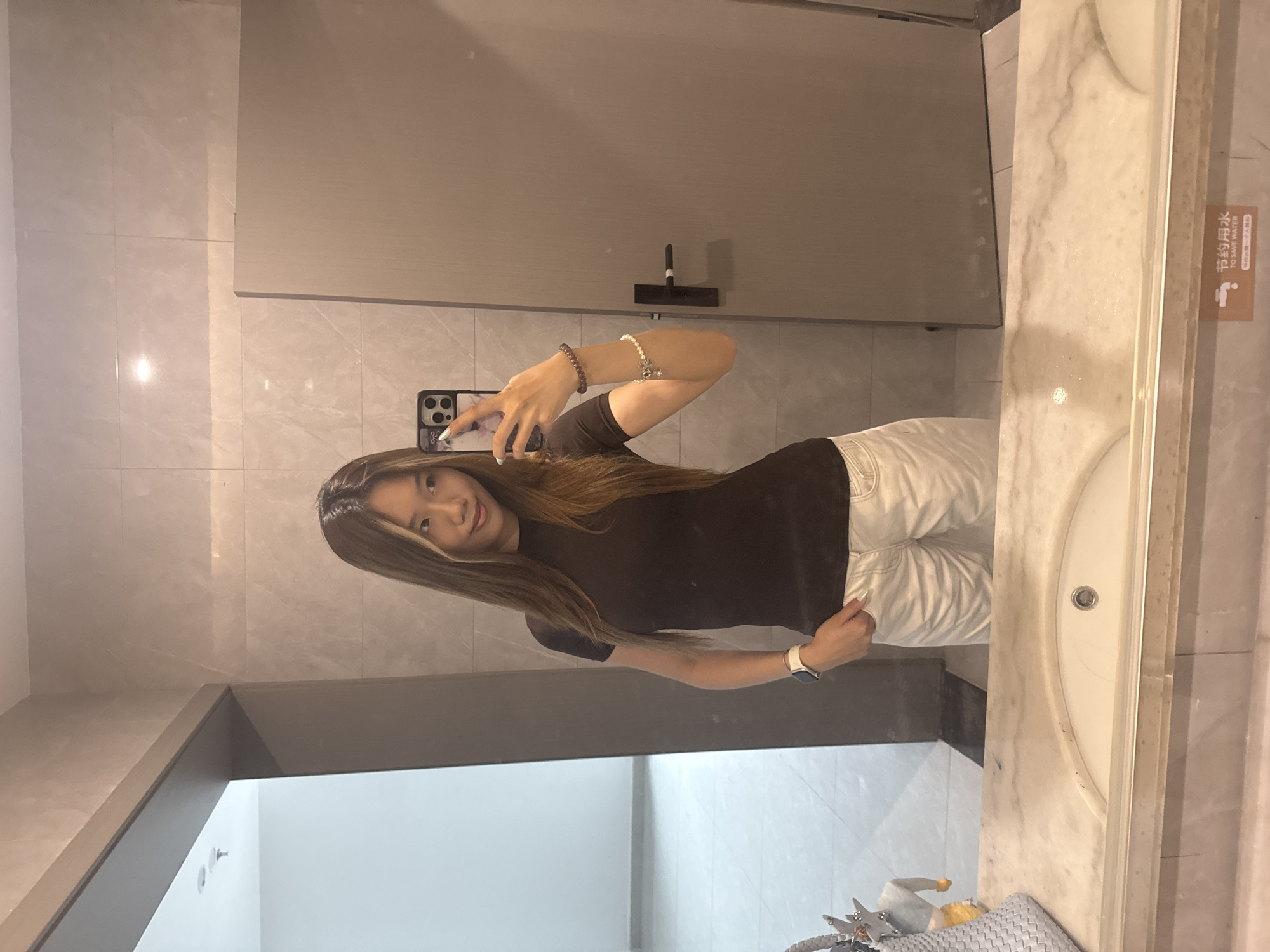 A young woman is taking a selfie in a bathroom. She is wearing a dark brown t-shirt and white pants. The bathroom has light gray tiled walls and a marble countertop with a sink. A sign with Chinese characters is visible on the right side of the image. The location is identified as being in the Nan'an District, China. The bathroom appears clean and modern.