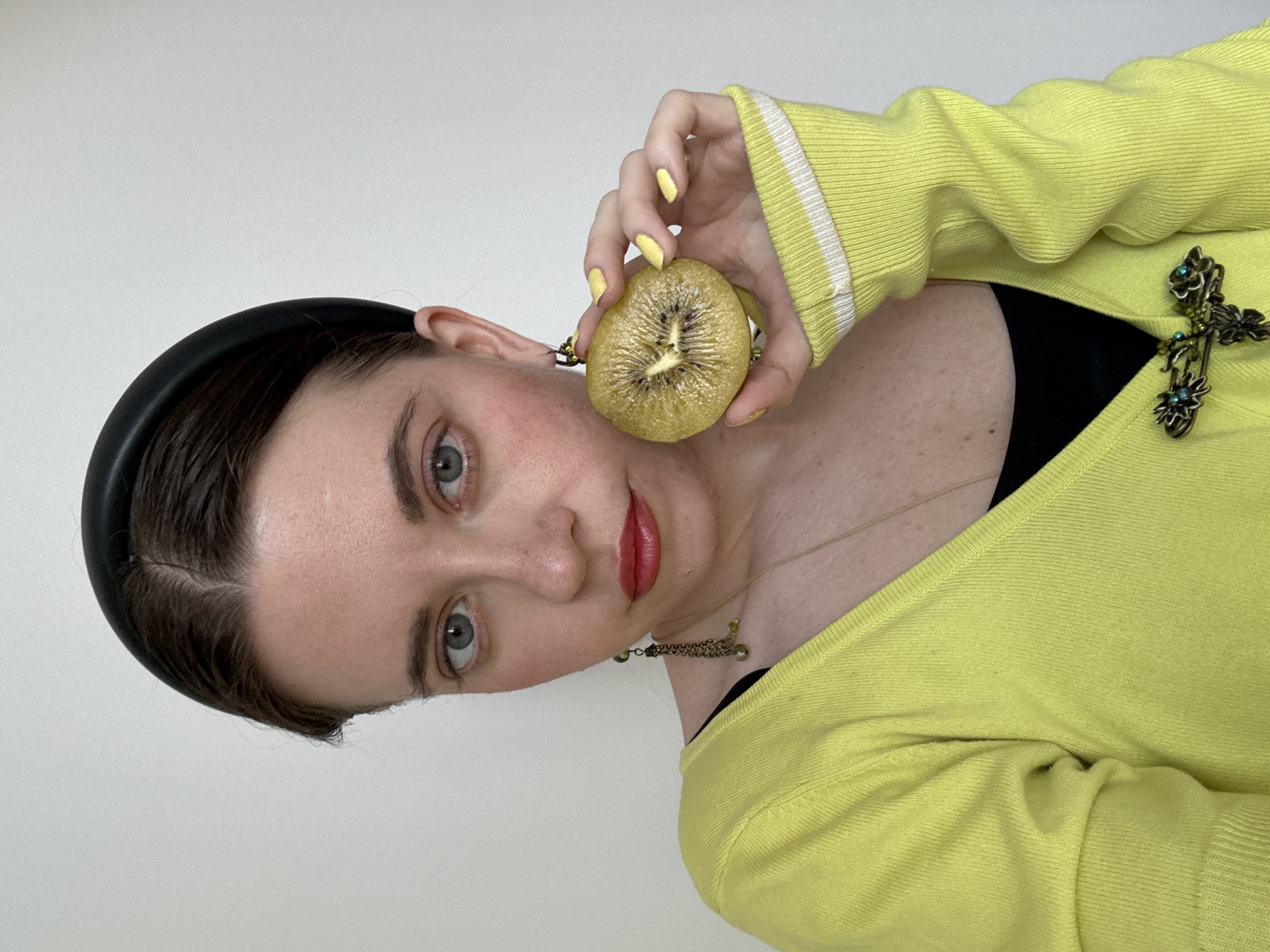 A person is holding a golden kiwi near their face. The person has a black headband, pale skin, and red lipstick. They are wearing a yellow sweater with a white stripe on the cuff and a cross-shaped brooch. A gold necklace is visible. The background is a plain white wall.