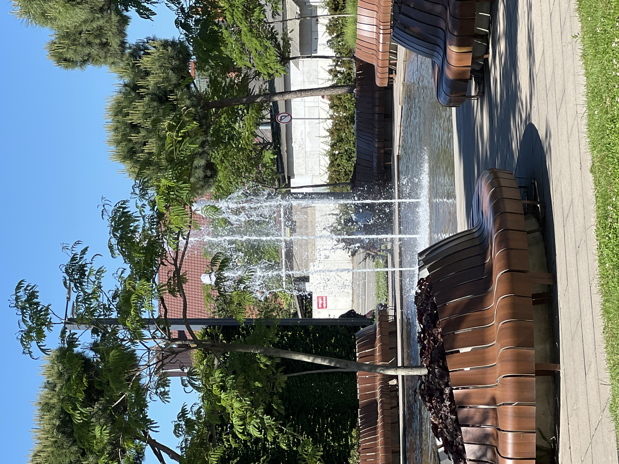A brightly lit image captures a vibrant public park scene in Krasnodar, Russia, under a clear blue sky. The central subject is a lively fountain, its jets of water spraying upwards, creating a refreshing atmosphere.
The scene features multiple modern, ergonomically designed wooden benches with a wave-like pattern, arranged along a paved walkway made of rectangular light-grey tiles. These benches line a narrow water feature, likely a canal or a decorative stream, from which the fountain emerges. To the right of the walkway, a strip of neatly maintained green grass is visible.
Various trees with lush green foliage are interspersed throughout the park. Some trees display distinctive light-colored, elongated blooms or cones, adding textural variety to the greenery. In the background, partially obscured by trees and the fountain's spray, are distant buildings, one with a reddish-brown tiled roof.
The strong, direct sunlight suggests it is mid-day, casting defined shadows from the benches and trees onto the paved path. No people are visible, lending a serene and tranquil feel to the park. A notable detail is a red circular traffic sign, partially visible amidst the trees and water, depicting a white horizontal bar with a left-pointing arrow underneath, indicating a "No Left Turn" or similar directional restriction on a nearby road. The overall setting is one of a well-maintained, contemporary urban green space.