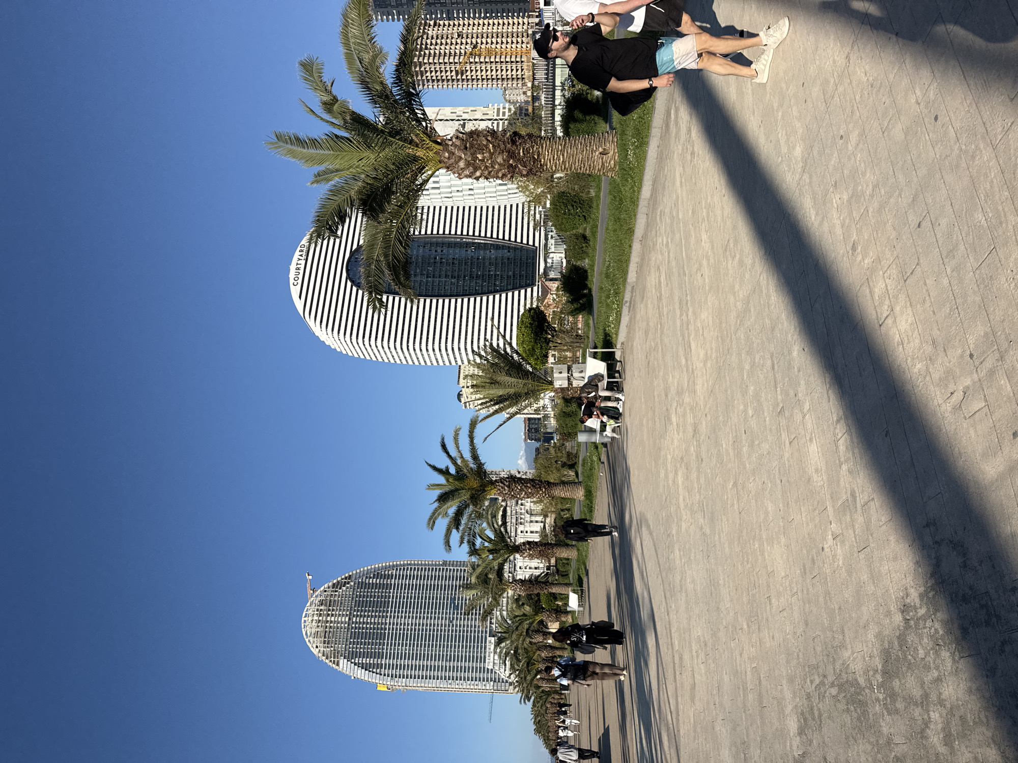 The image shows a pedestrian promenade in Batumi, Georgia, with modern architectural buildings and palm trees lining the walkway. The Courtyard by Marriott hotel, with its distinctive white and black striped facade, is prominent in the scene. Further down the promenade, a taller, elliptical building is visible. The sky is clear and blue. A group of people are walking on the right-hand side of the image, including a person wearing a black shirt, light blue shorts, and a baseball cap. The promenade surface appears to be concrete.