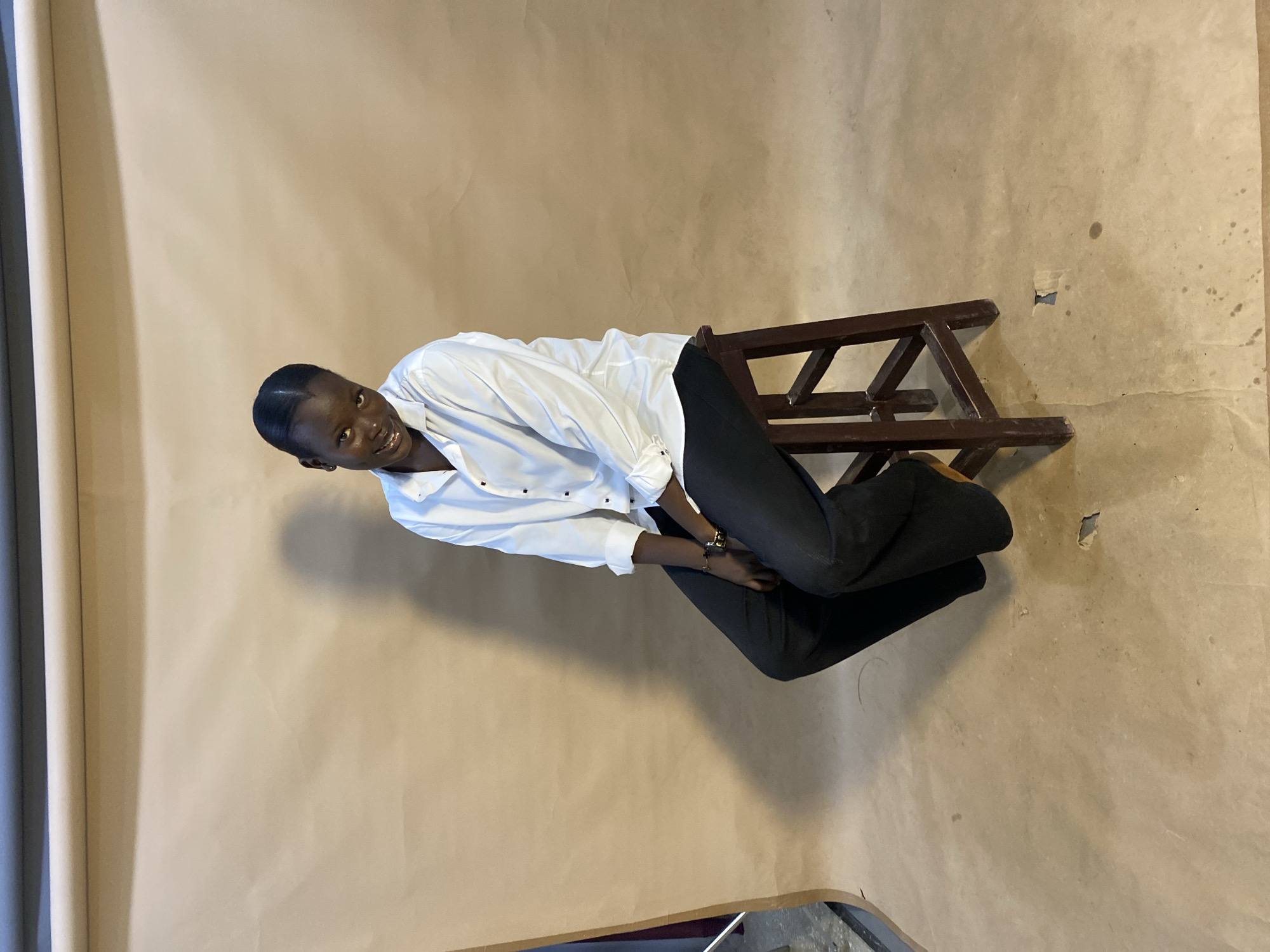 A young Black person is seated on a dark brown wooden stool against a plain, light brown backdrop. The person is wearing a white long-sleeved button-down shirt with the sleeves rolled up and black wide-leg trousers. They are looking directly at the camera with a slight smile and their head is tilted to their left. Their dark hair is pulled back. The lighting casts a shadow of the person on the backdrop to their left. The background appears to be a studio setting with a large paper roll at the top. The floor is also the same light brown paper material. The image was taken in Jos, Nigeria.