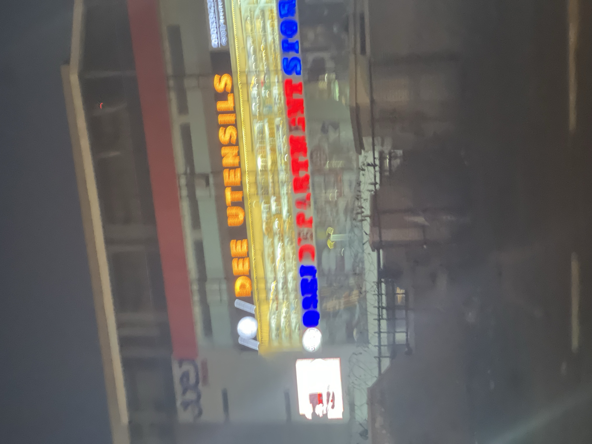 This media file captures a multi-story commercial building in Abuja, Nigeria, at night or dusk. The primary subjects are two brightly illuminated signs on the building's facade. The upper sign features orange and yellow block letters, clearly spelling out "DEE UTENSILS". Below it, a second sign uses red and blue lettering, which reads "OSRI DEPARTMENT STORE" (with "STOR" visible, implying "STORE").
The scene is set in an urban environment, characterized by the dark sky and the vibrant glow of the neon signs, indicating the late hours. The building itself has a notable red horizontal stripe running across its structure. Architectural details of the building and its immediate surroundings, including what appears to be security fencing or railings in front, are somewhat blurred due to the low light conditions and possibly motion, but the signs remain sharp. A tiny, crescent-shaped red light is faintly visible on the upper left section of the building. There are no people visible, and no specific activity is taking place, beyond the implied commercial operations of the stores. The overall impression is of a lively business district within Abuja after sunset.