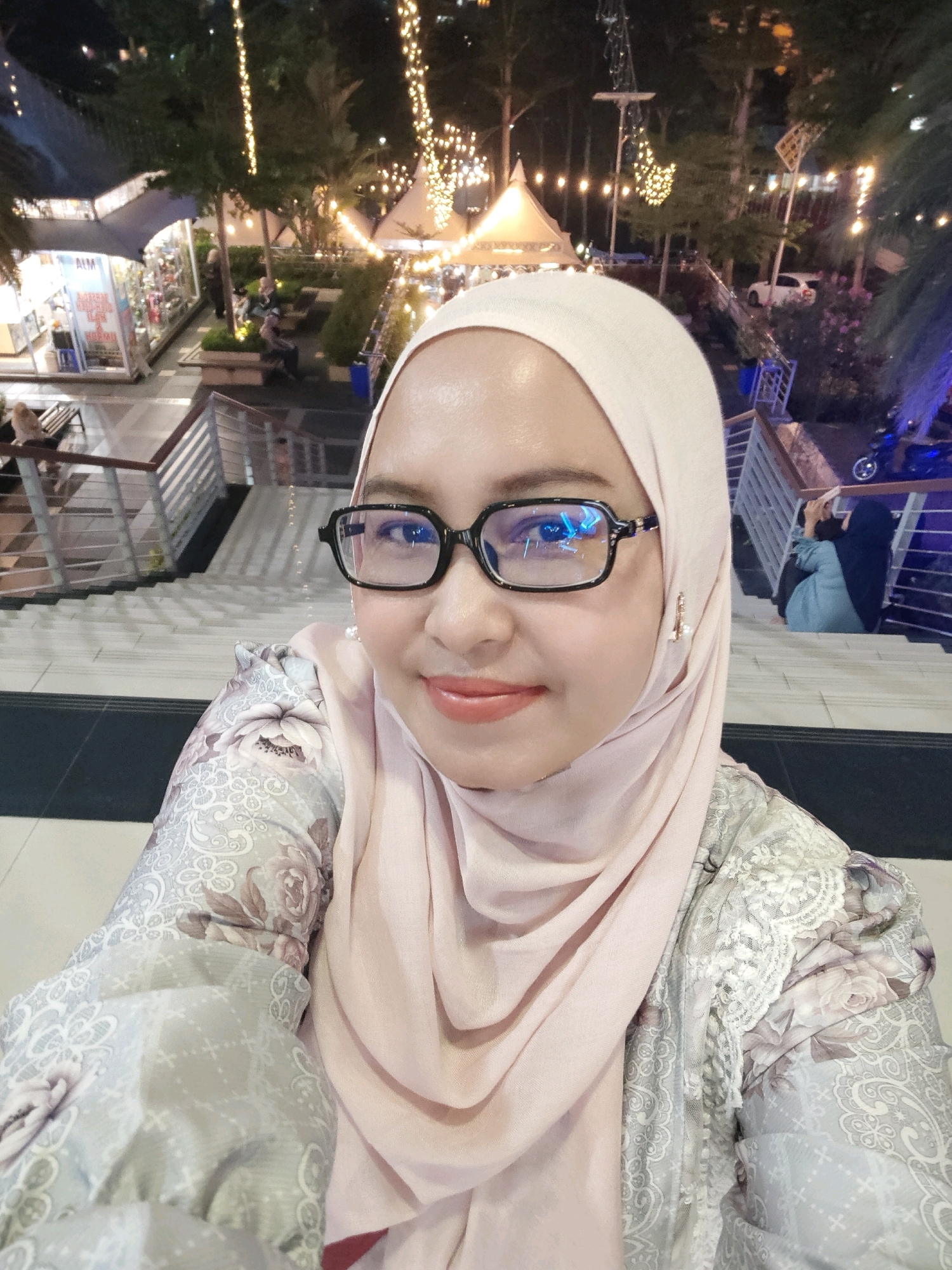 The image is a selfie of a woman wearing a light pink hijab and glasses. She is smiling and looking at the camera. She is wearing a light-colored shirt with a floral pattern. In the background, there are lights strung between trees, tents, and stairs. There are also people walking and sitting in the background. The scene appears to be an outdoor market or festival at night. The woman's attire suggests she is likely Indonesian, and the general ambiance and architecture point to a location like Nagoya Hill Shopping Mall in Batam, Indonesia, a popular spot for evening strolls and events.