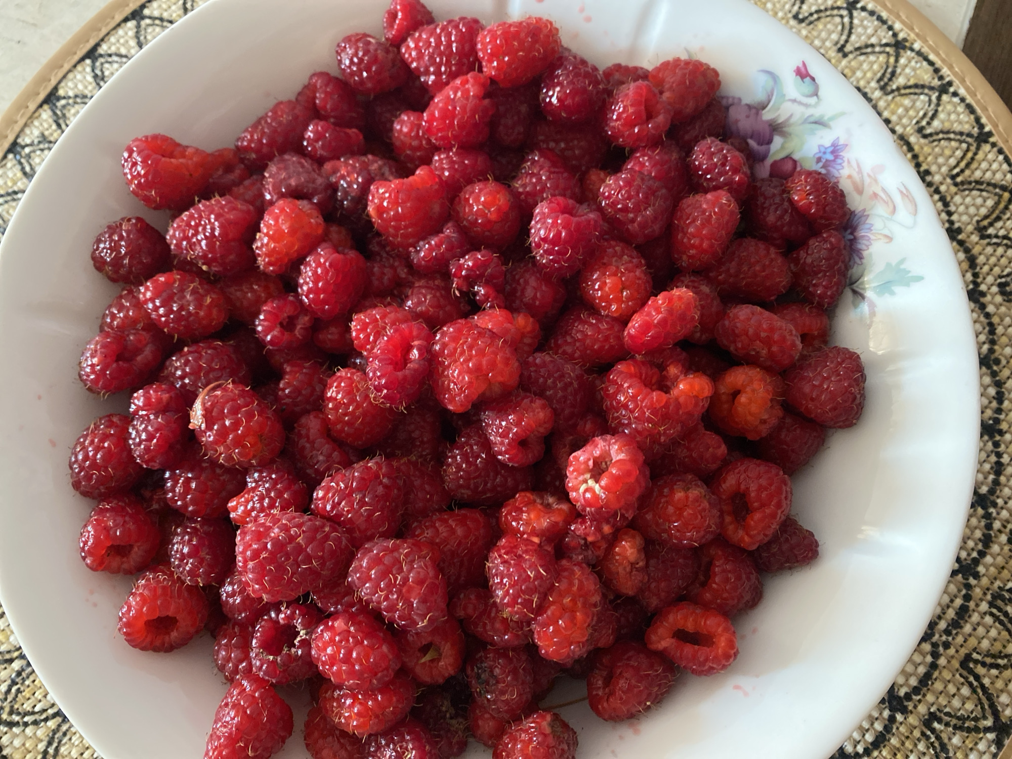 A close-up, overhead shot shows a white ceramic bowl filled with fresh raspberries. The bowl has a floral pattern around its rim, featuring green leaves and purple and pink flowers. The raspberries are a deep red color, with some appearing slightly darker or lighter than others. They are piled high in the bowl, with many individual drupelets visible. The bowl rests on a textured placemat with a woven pattern in shades of beige and brown. The background is slightly blurred, but appears to be a light-colored surface. There are no people or animals visible in the image. The setting is likely a domestic environment, possibly a kitchen or dining area. There is no information in the image that allows for the specific location within the city of Lida, Belarus to be identified.