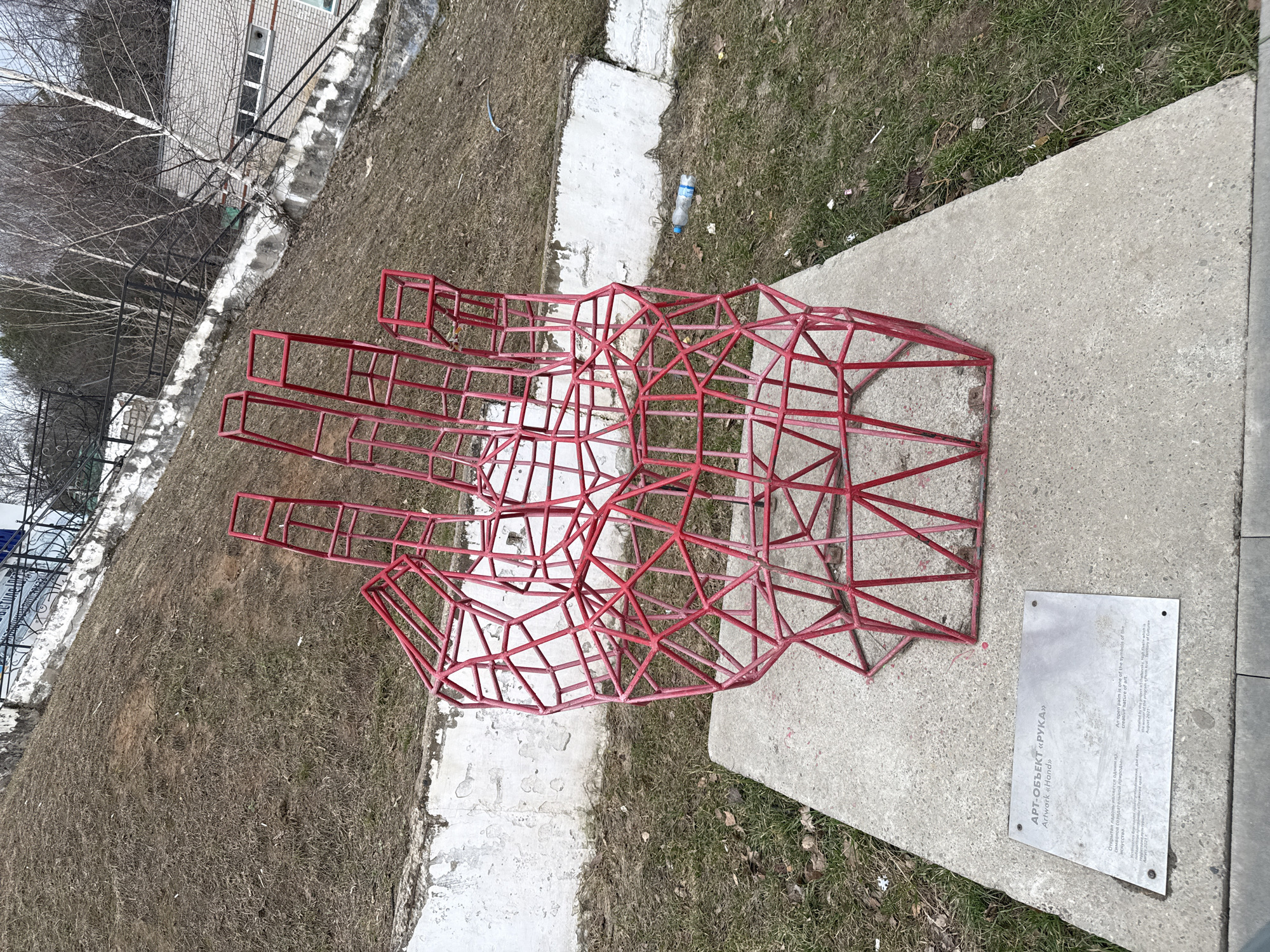 Here's a detailed description of the image:
**General Scene:** The image presents an outdoor urban setting with a red, wireframe sculpture of a hand as the central subject. The sculpture is situated on a concrete slab, and there's a plaque attached to the slab, presumably providing information about the artwork. The surroundings include a grassy area with some bare patches, indicating a possibly early spring or late fall season.
**Objects & Subjects:**
* **Red Wireframe Hand:** The most prominent object is the sculpture of a hand constructed from red-painted metal rods. The rods form a geometric, open structure, outlining the shape of a human hand with visible fingers and palm.
* **Concrete Slab:** The hand sculpture rests on a rectangular concrete slab, which appears to be its base.
* **Plaque:** A metal plaque is mounted on the concrete slab near the sculpture. It likely contains the title of the artwork ("АРТ-ОБЪЕКТ «РУКА»" or "Artwork 'Hand'") and other relevant information.
* **Surrounding Environment:** The area around the concrete slab consists of a patch of grass, partially dry or dormant, with some bare earth visible. A few scattered dry leaves are present on the ground. There is also a plastic bottle in the background.
* **Background:** The background features a building with a window, and some bare trees and a fence.
**Environment:**
* **Location:** The photograph appears to be taken in a park or open area within a city. The specific location (Unknown, Unknown) is provided.
* **Weather/Season:** The scene suggests either early spring or late fall.